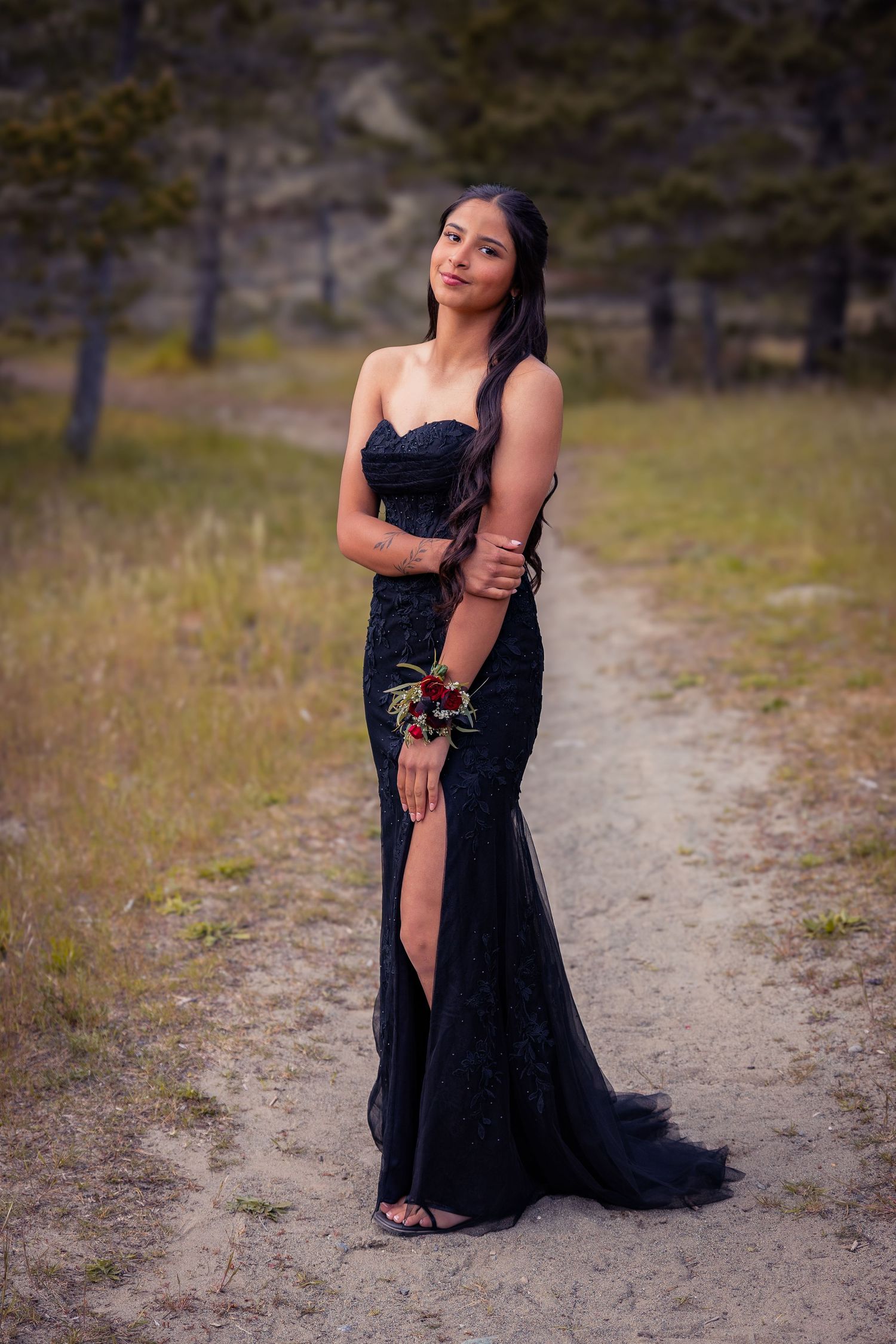 Young woman in elegant black strapless mermaid prom dress with floral wrist corsage posing on a dirt path outdoors.