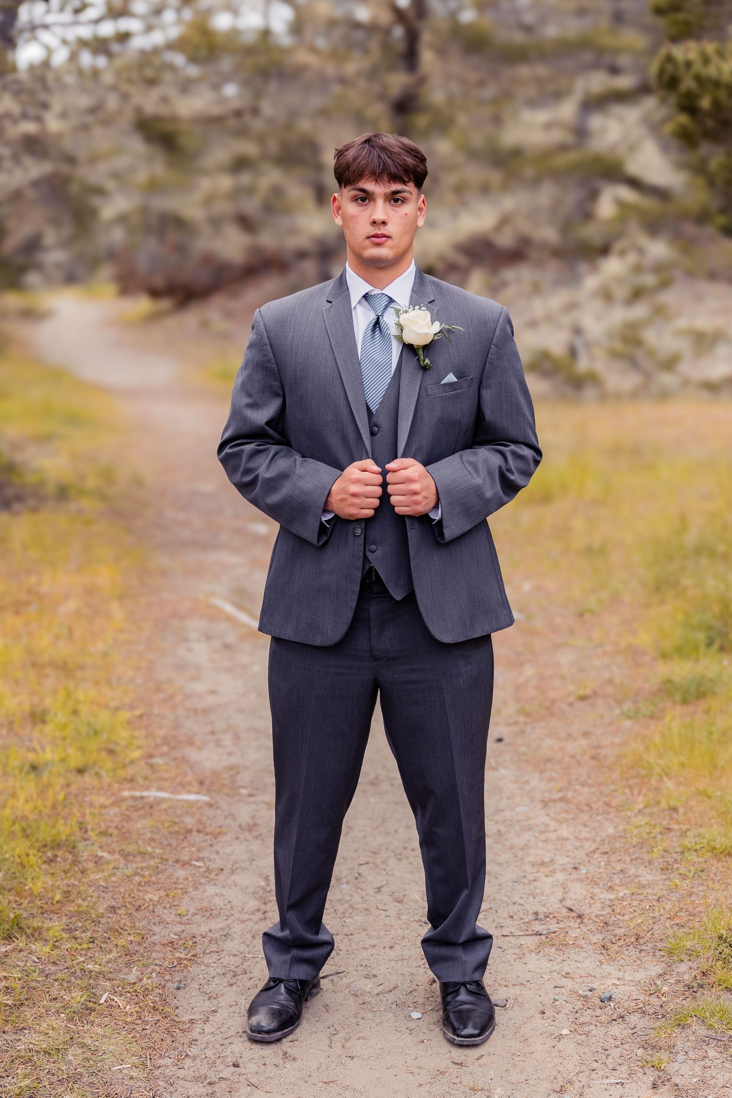Young man in gray suit with blue tie and white boutonniere stands on dirt path for formal portrait.