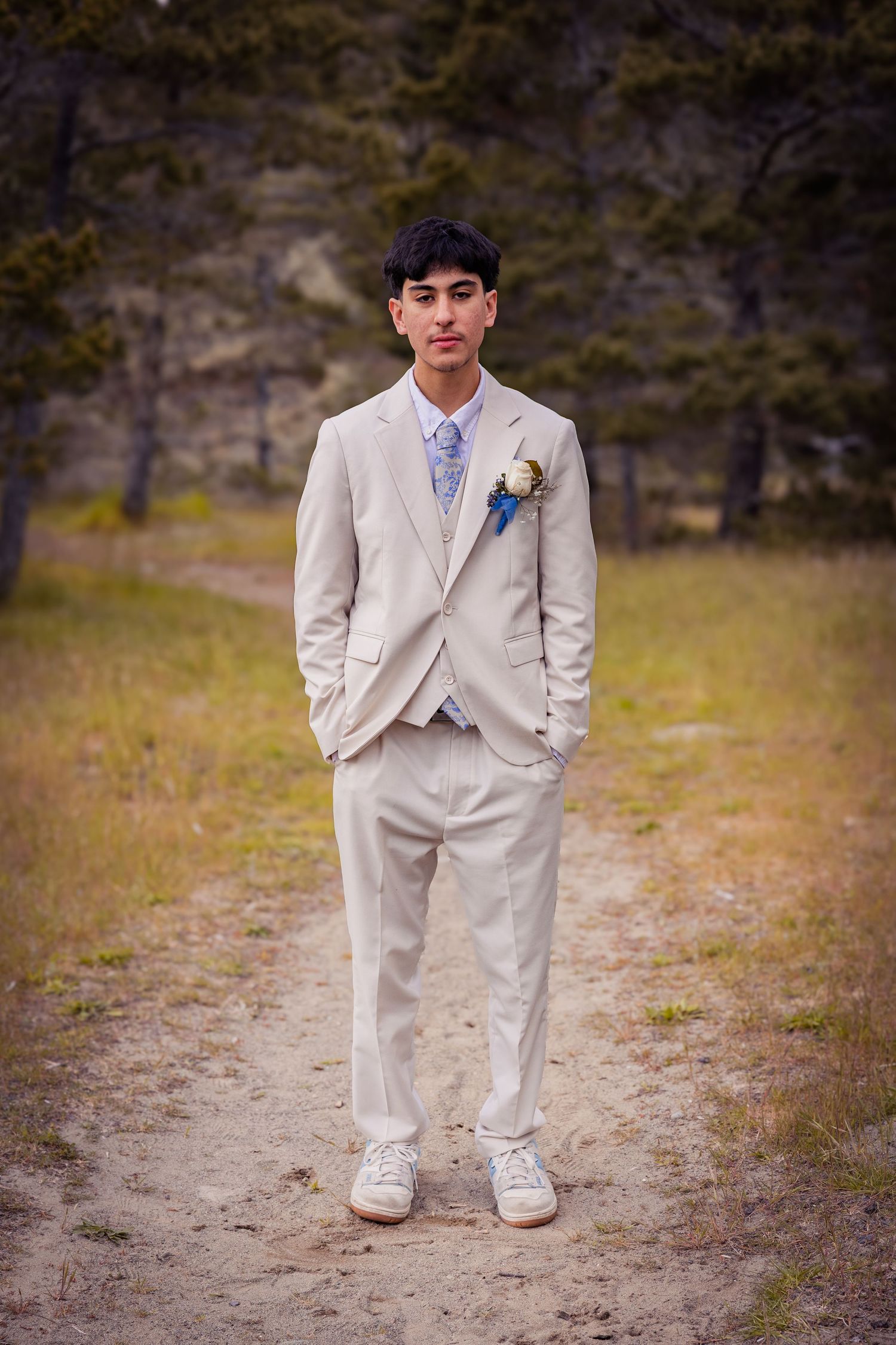 Young man in beige suit with boutonniere and blue tie stands on sandy path surrounded by trees.