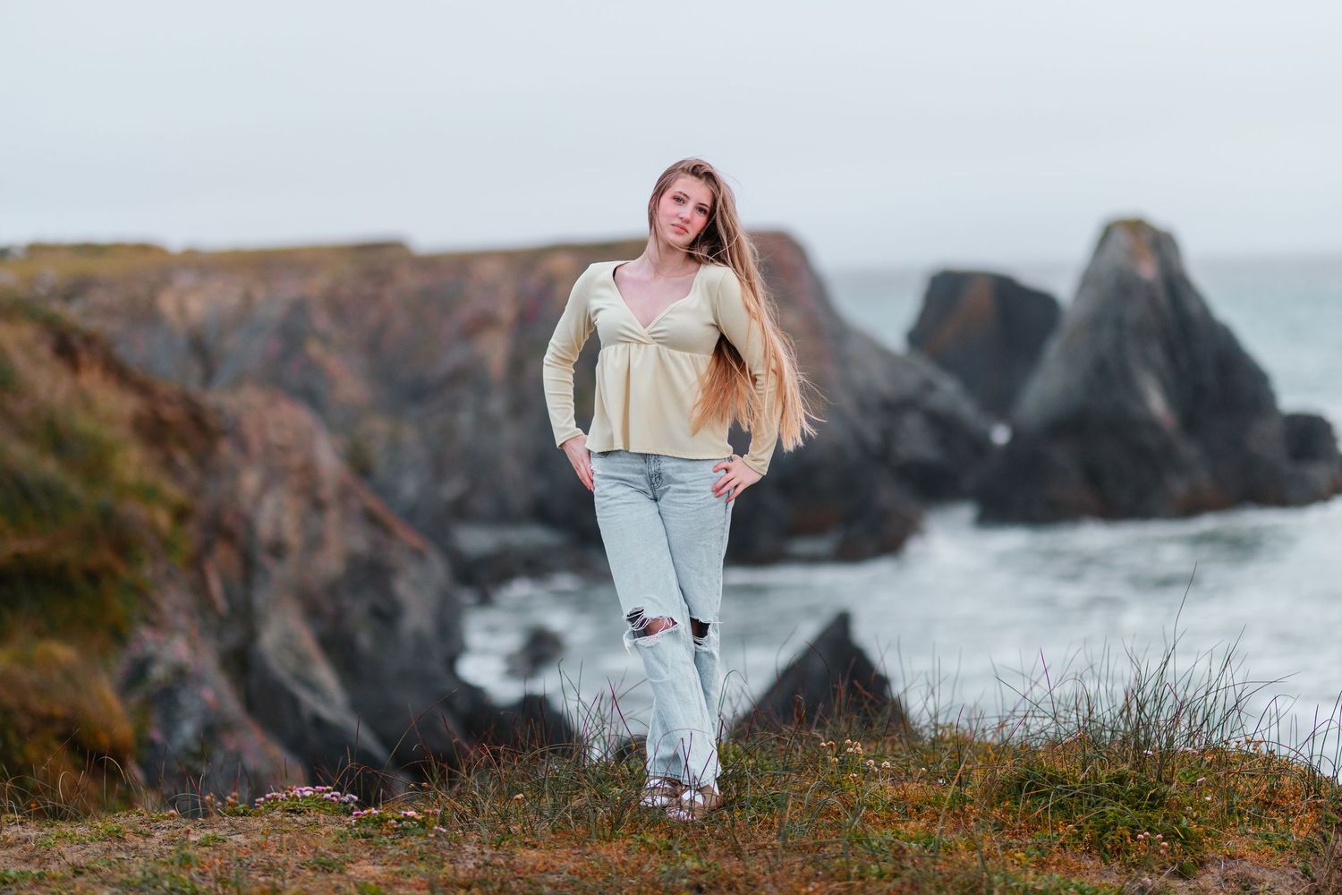 Young woman with long blonde hair poses in ripped jeans on rocky coastal cliffs with ocean waves in background.