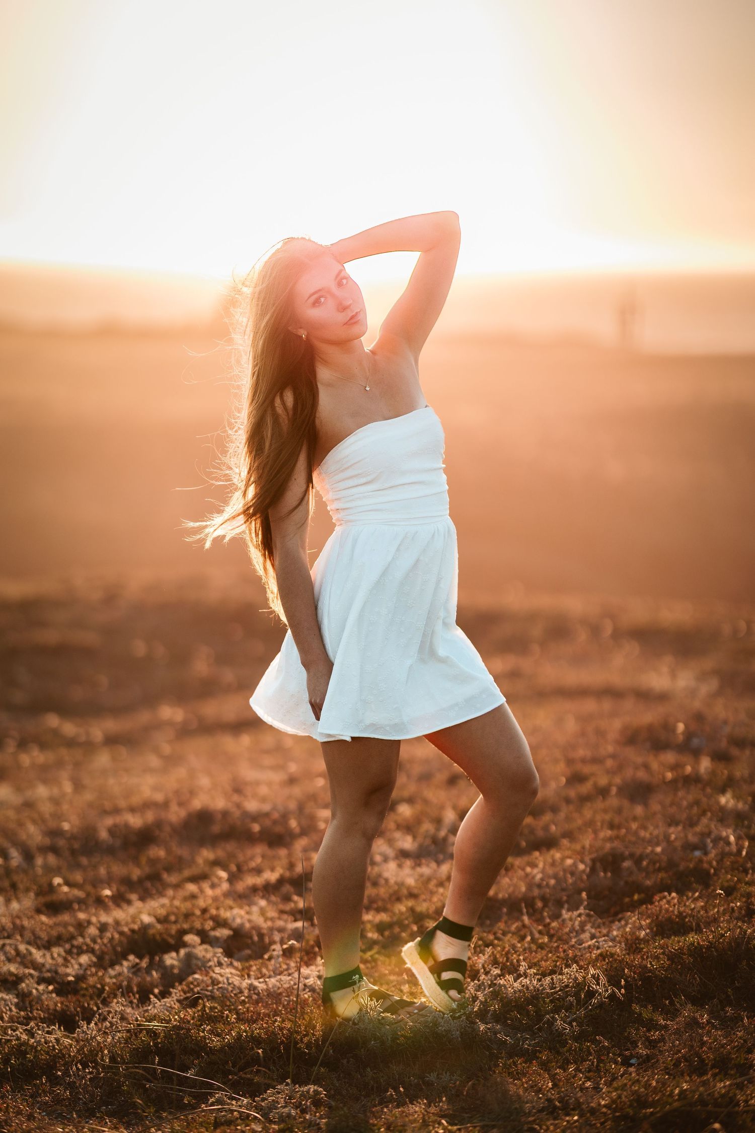 Young woman in white dress dances in open field at golden hour with hair flowing in the wind.