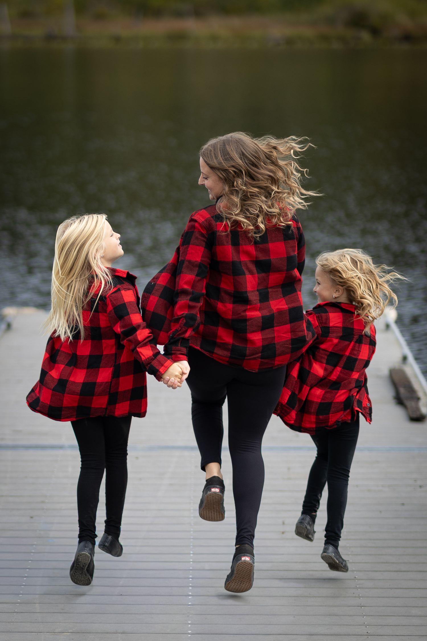 A mother and two daughters in matching red plaid shirts hold hands on a dock by a lake.