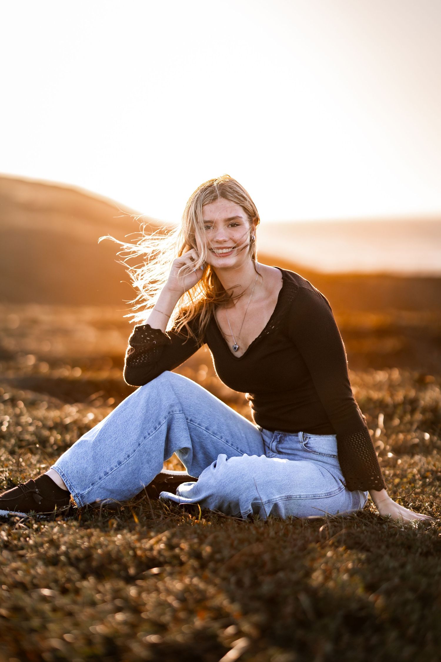 Smiling blonde teen girl sitting in golden field at sunset wearing black top and light blue jeans.