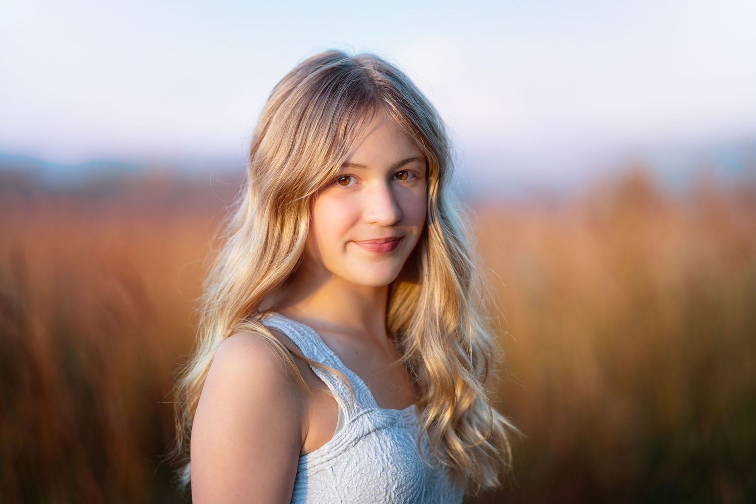 Blonde teenage girl in white dress smiling softly outdoors in warm golden hour light.