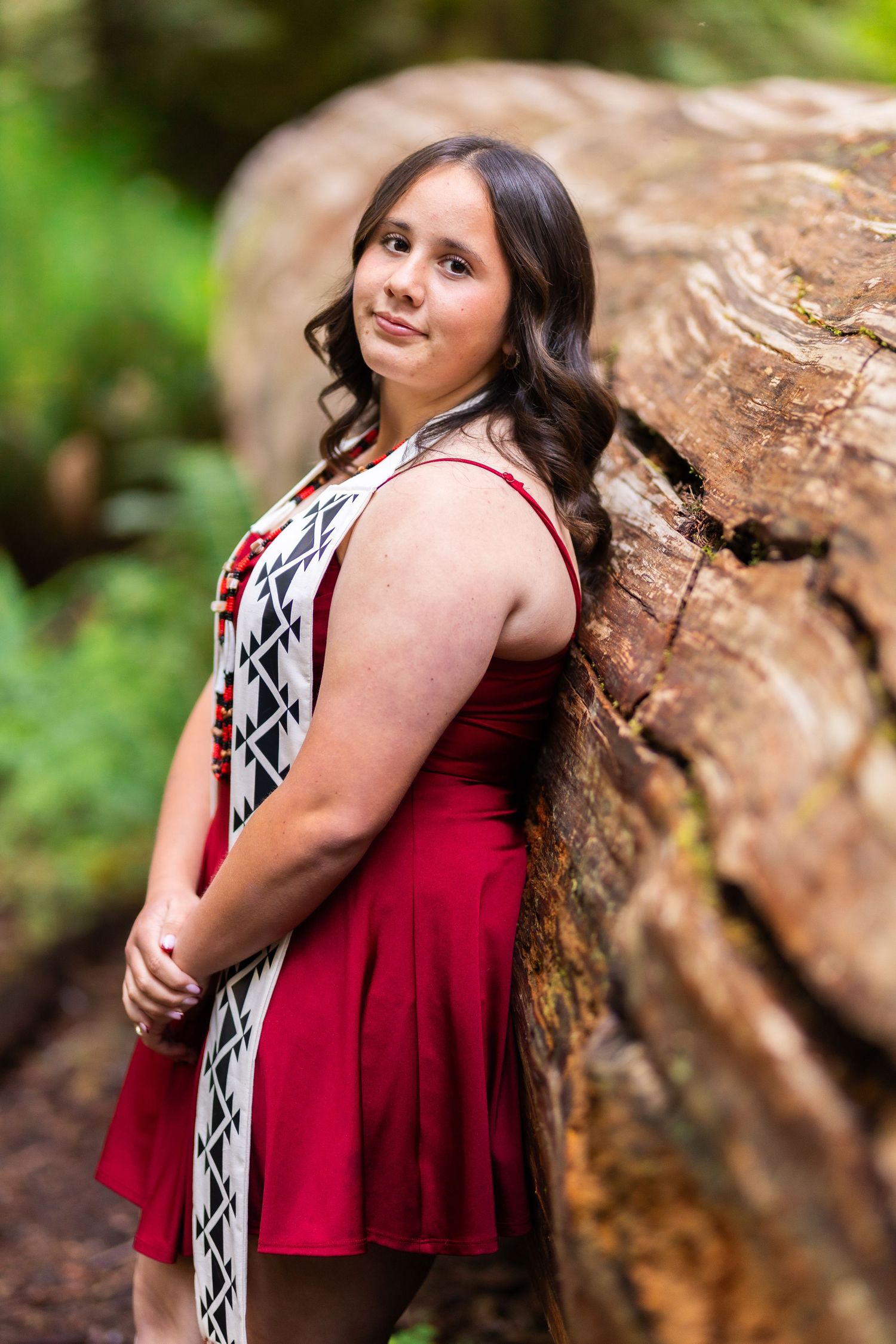 Young woman in red and white dress posing against a large rock in a lush green forest setting.