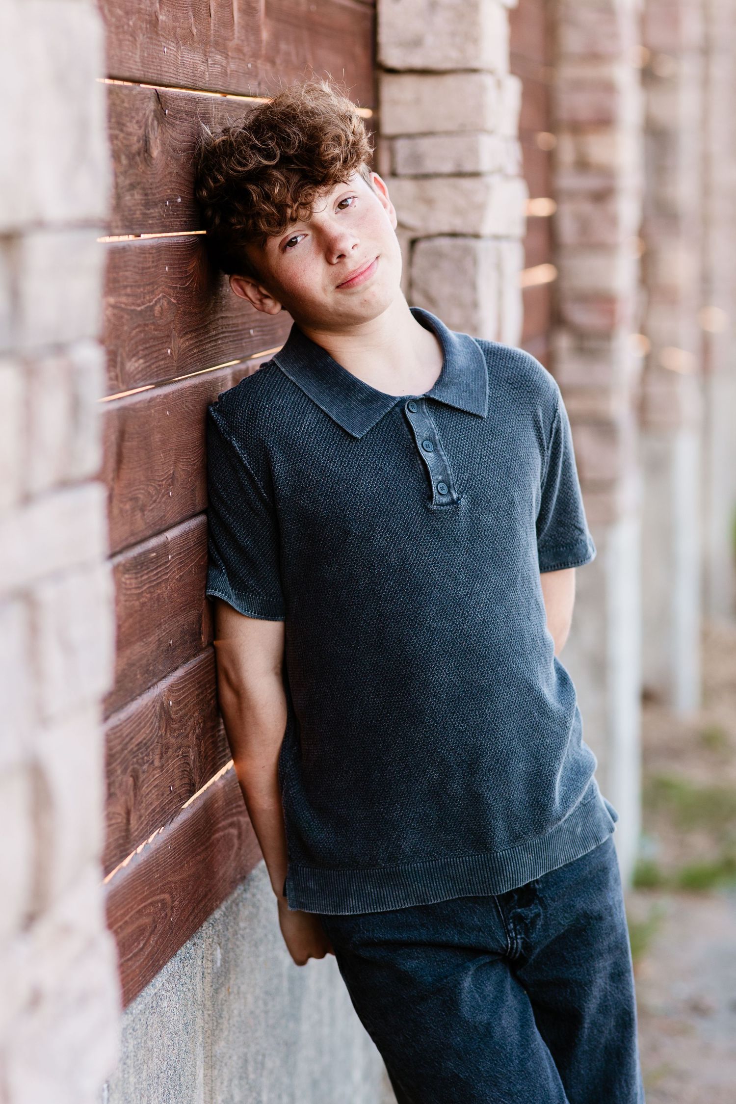 Teen boy with curly hair leans against brick wall wearing dark polo shirt and jeans, smiling casually.