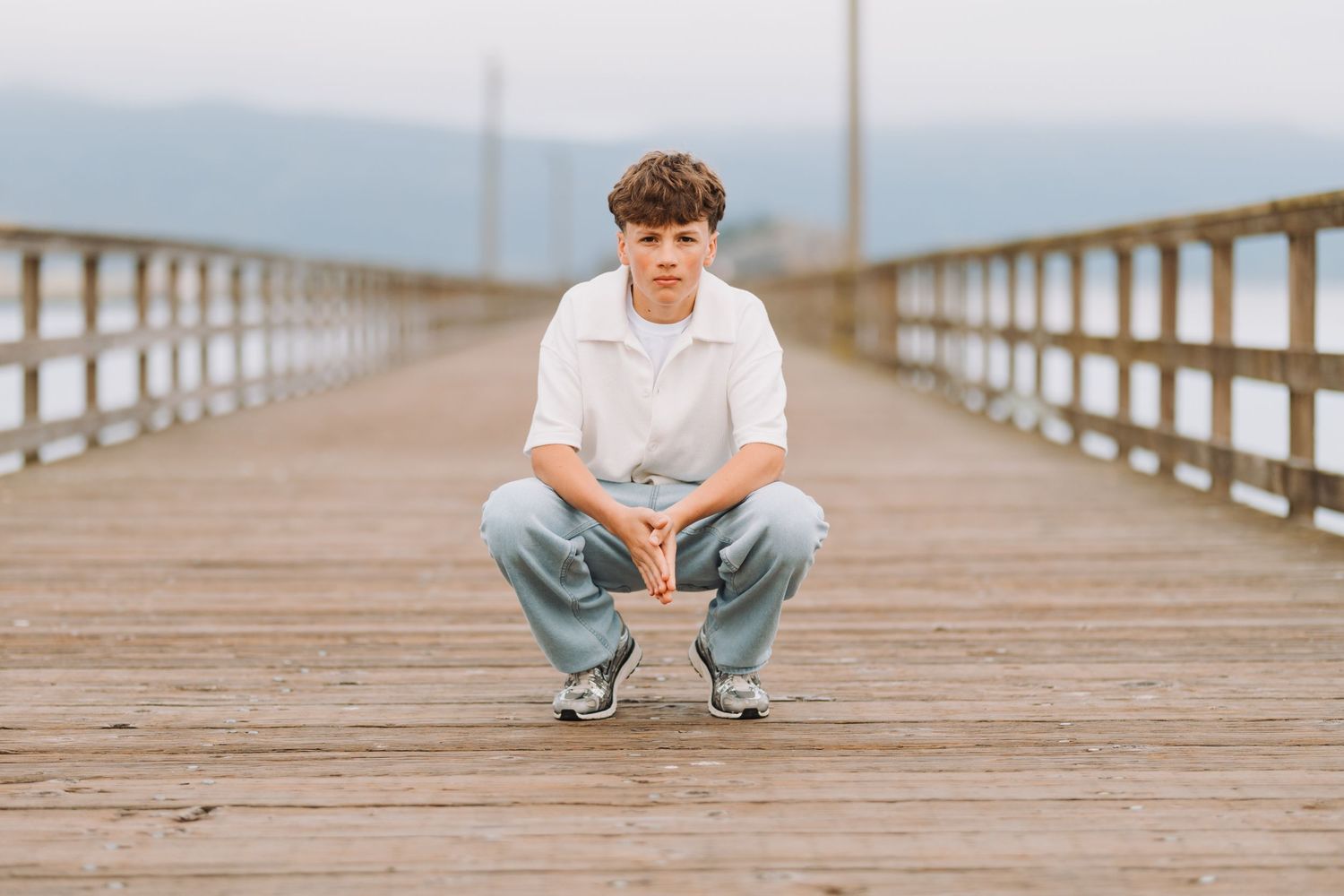 Young man crouching on a wooden pier with mountain backdrop, wearing white shirt and light jeans.