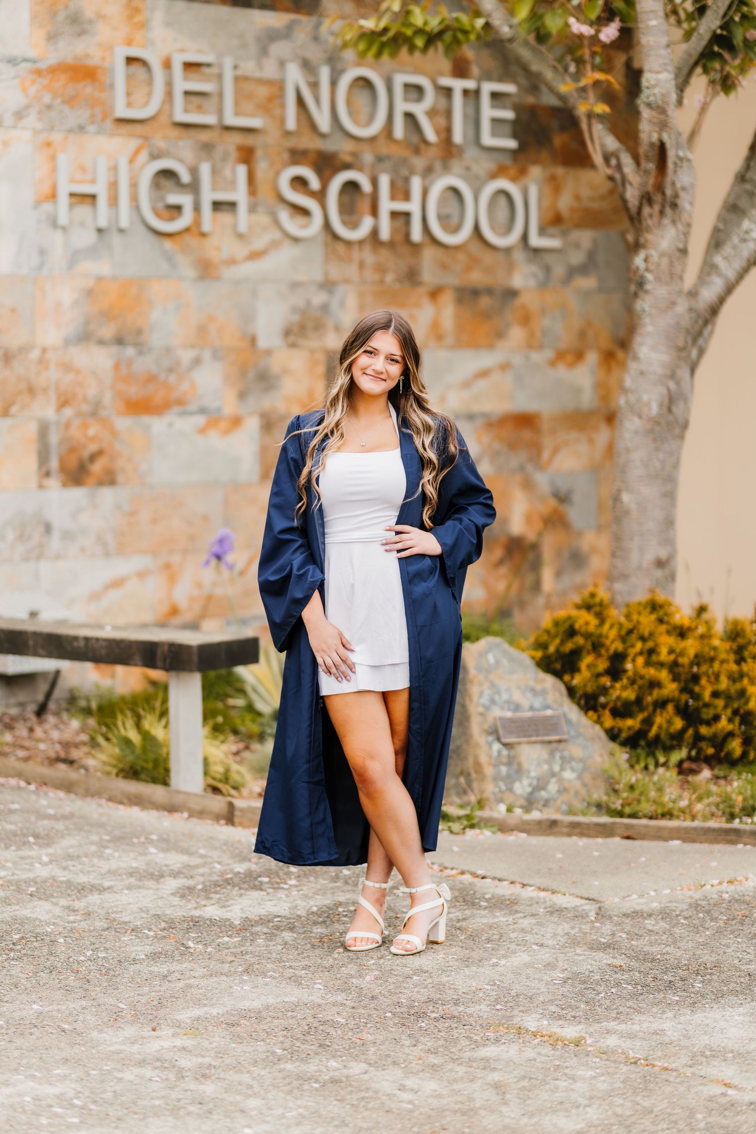 Senior girl in navy graduation gown poses in front of Del Norte High School stone sign wearing white dress and heels.