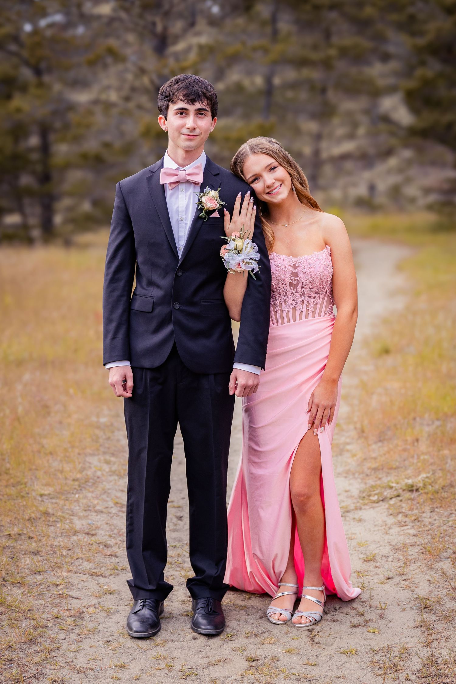 A teenage couple dressed in prom attire, boy in black suit with pink bow tie, girl in pink gown, posing outdoors.