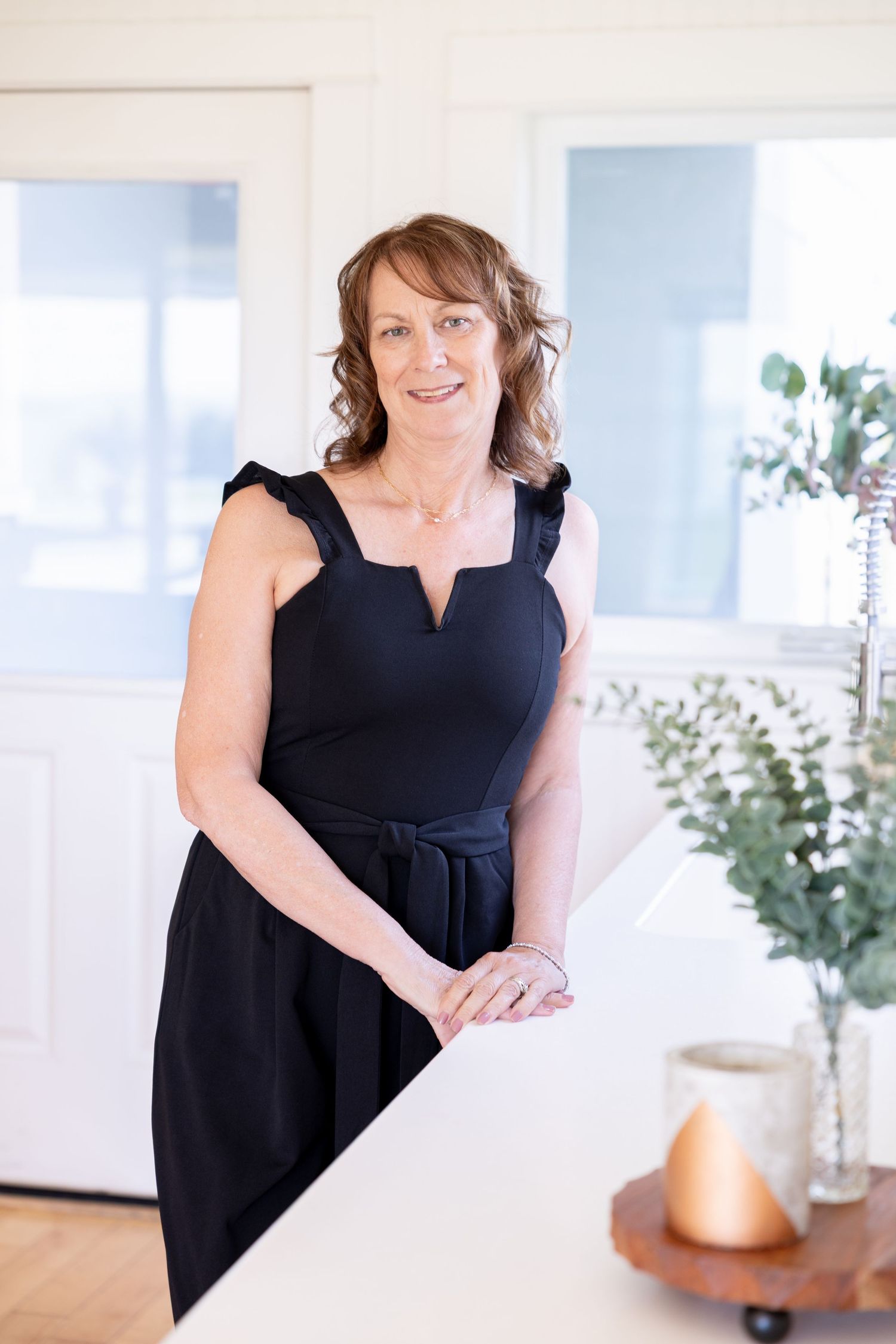 A smiling middle-aged woman in a black sleeveless dress leans against a white counter in a bright, modern interior.