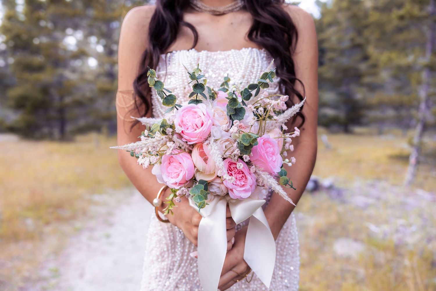 Bride in lace dress holding a pink peony and eucalyptus wedding bouquet with white ribbon outdoors.