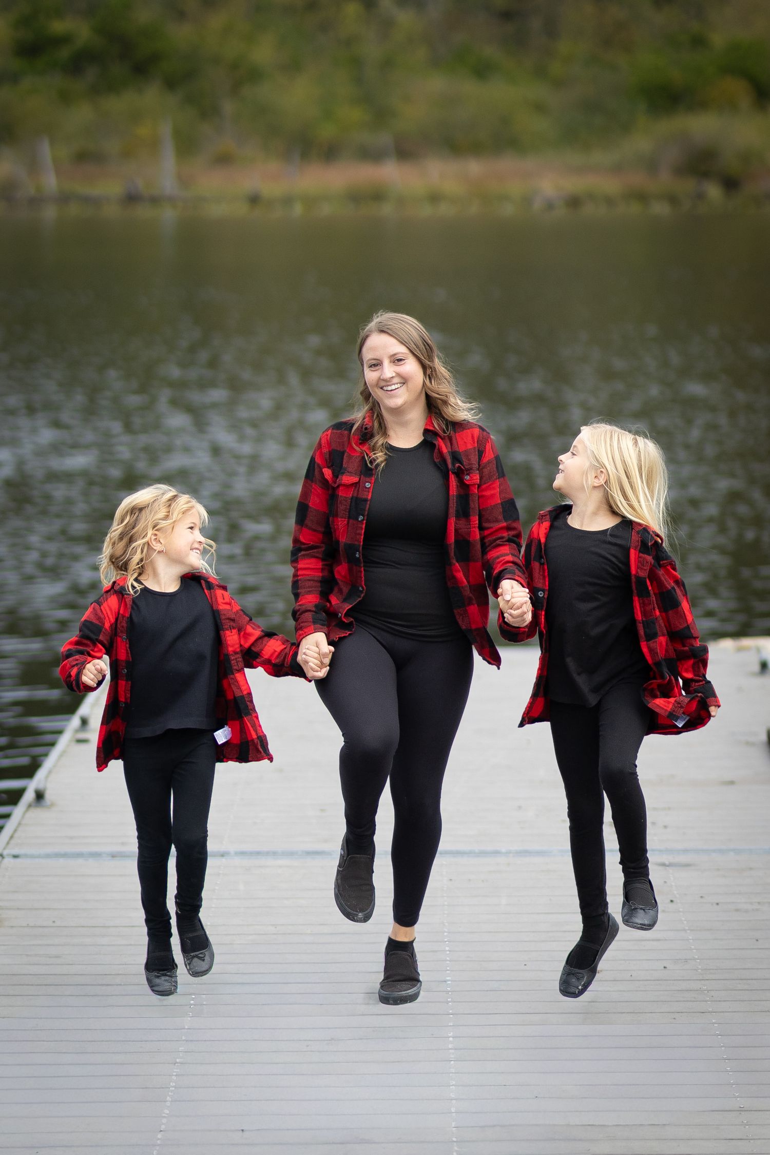 Mother and two daughters in matching red plaid shirts walk hand-in-hand on a dock by a lake.