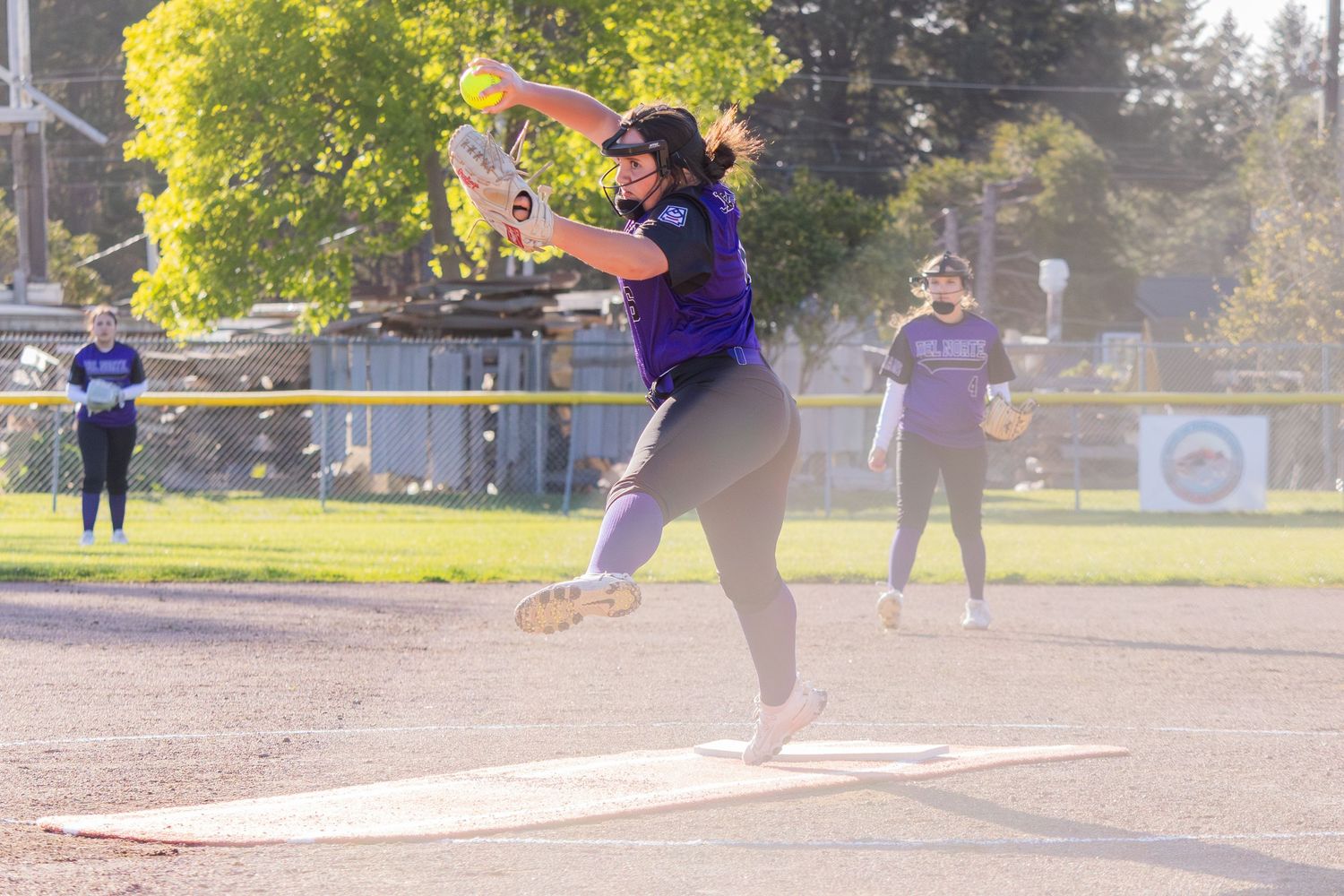 Softball pitcher in purple uniform winds up for a powerful pitch during an outdoor game in evening light.