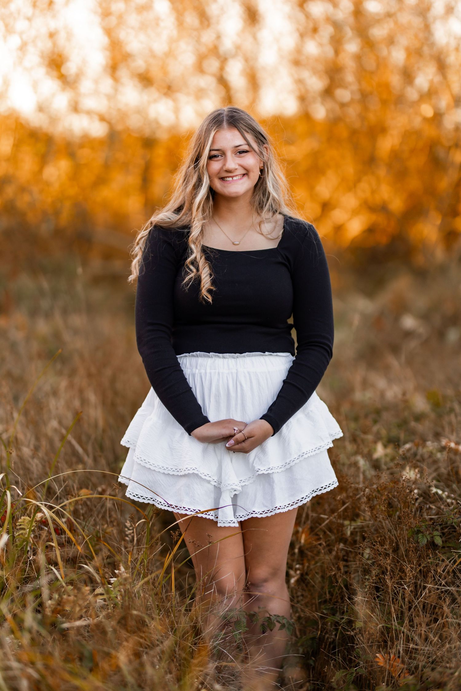 Smiling blonde teen girl in black top and white ruffle skirt posing in autumn field with golden foliage.