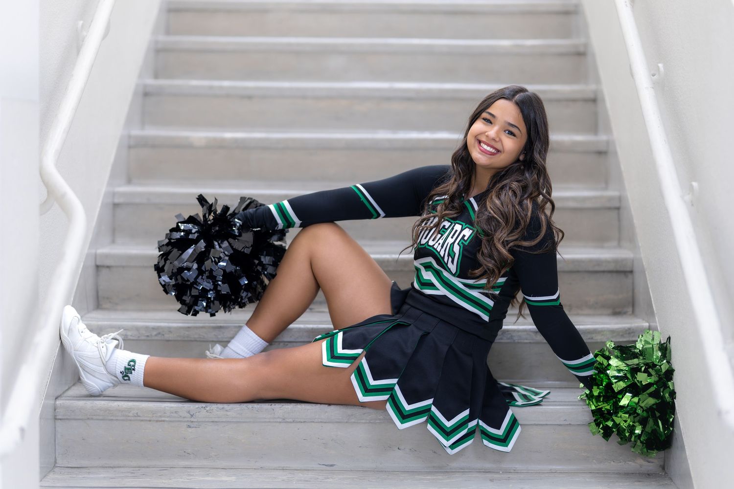 A cheerleader in a black and green uniform sits on stairs holding pom-poms and smiling.