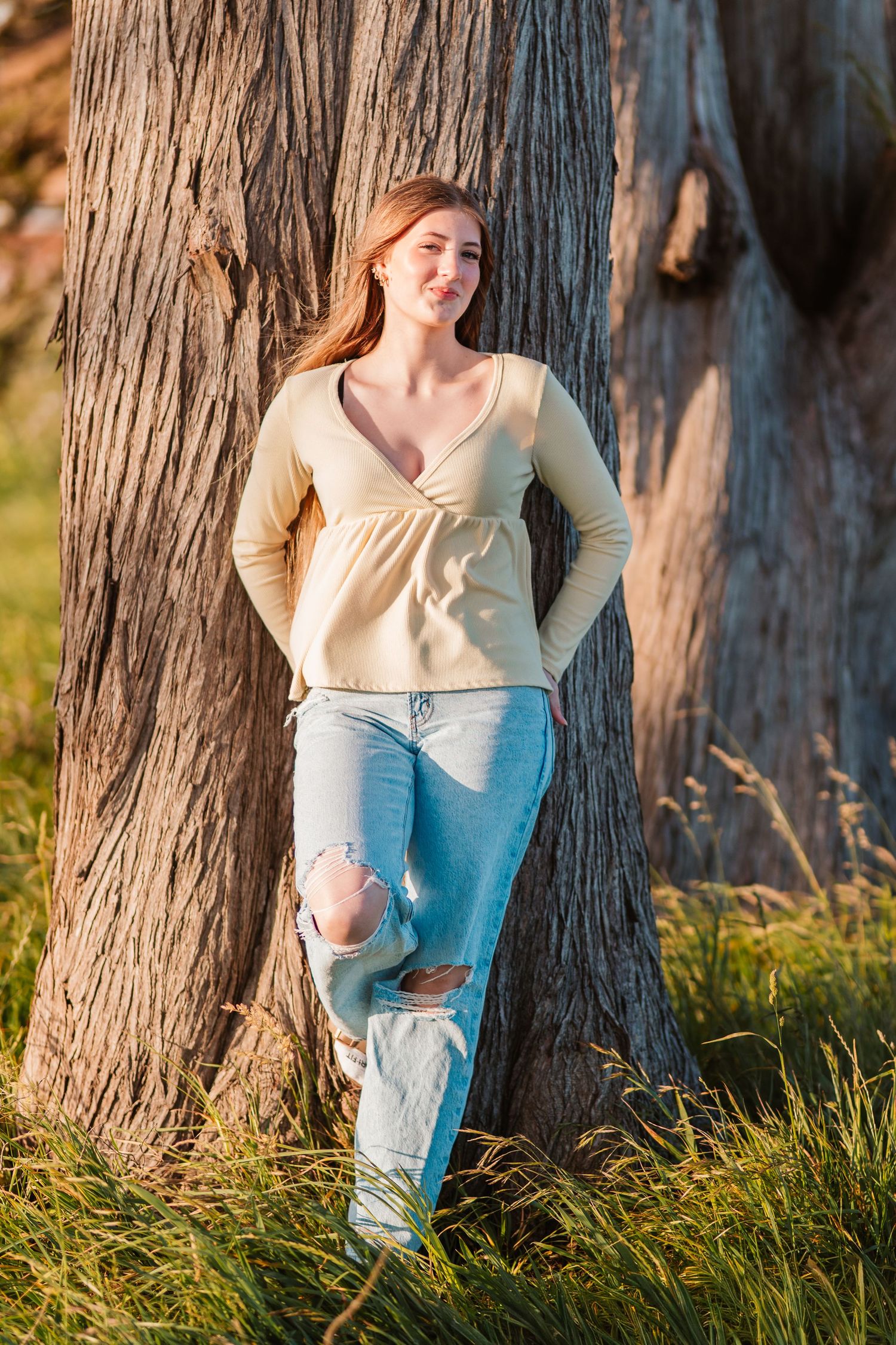 Red-haired teen girl in yellow top and ripped jeans leans against a large tree trunk in sunlit grass.