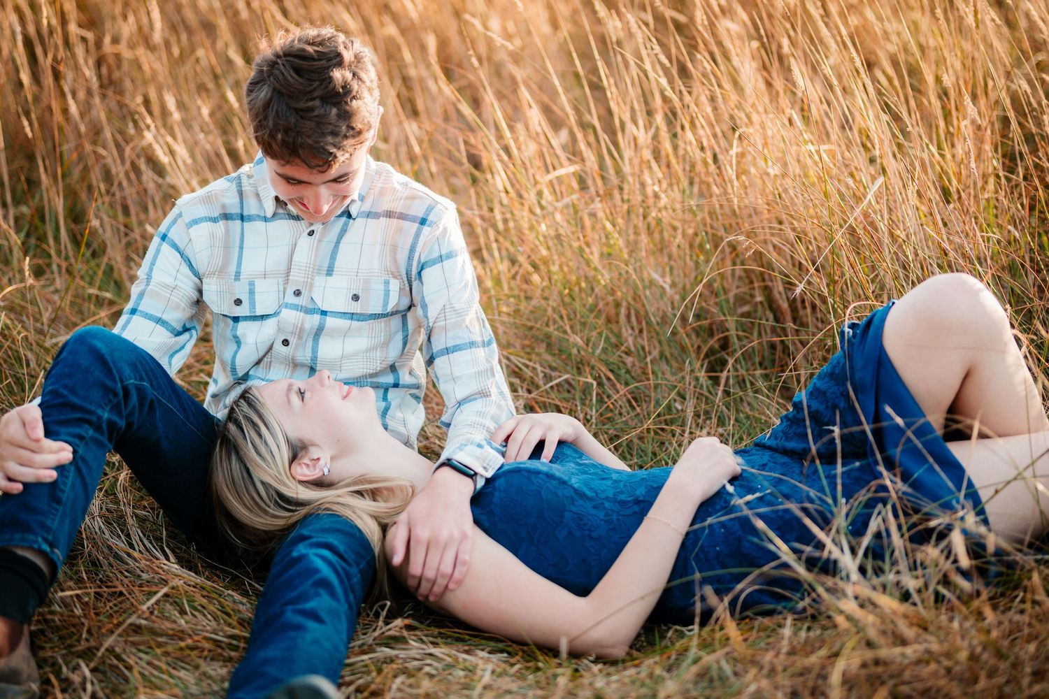 A couple relaxing in tall golden grass field, woman laying across mans lap in warm sunlight.