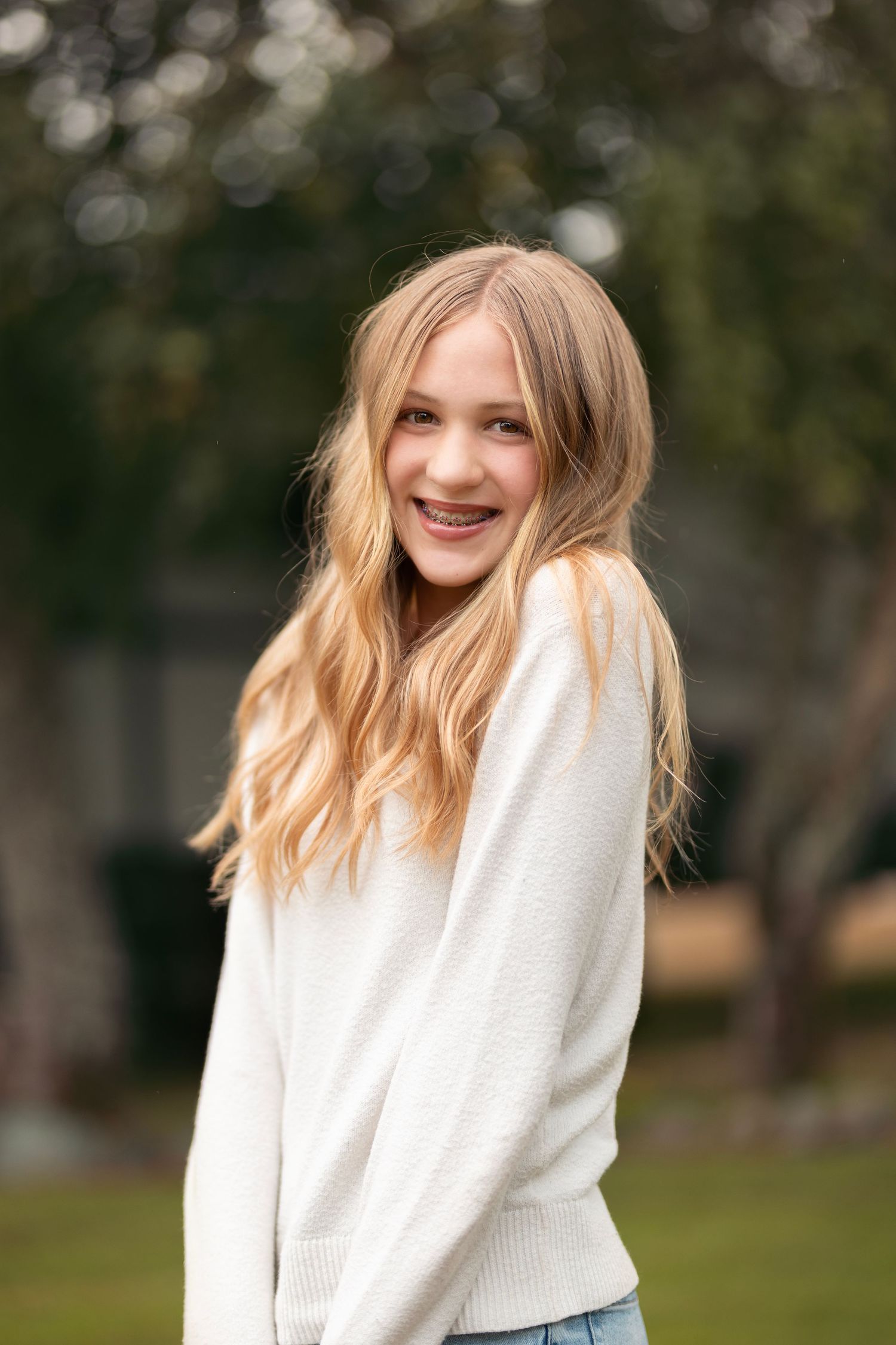 Smiling blonde teenage girl with braces in white sweater posing outdoors with green trees behind her.
