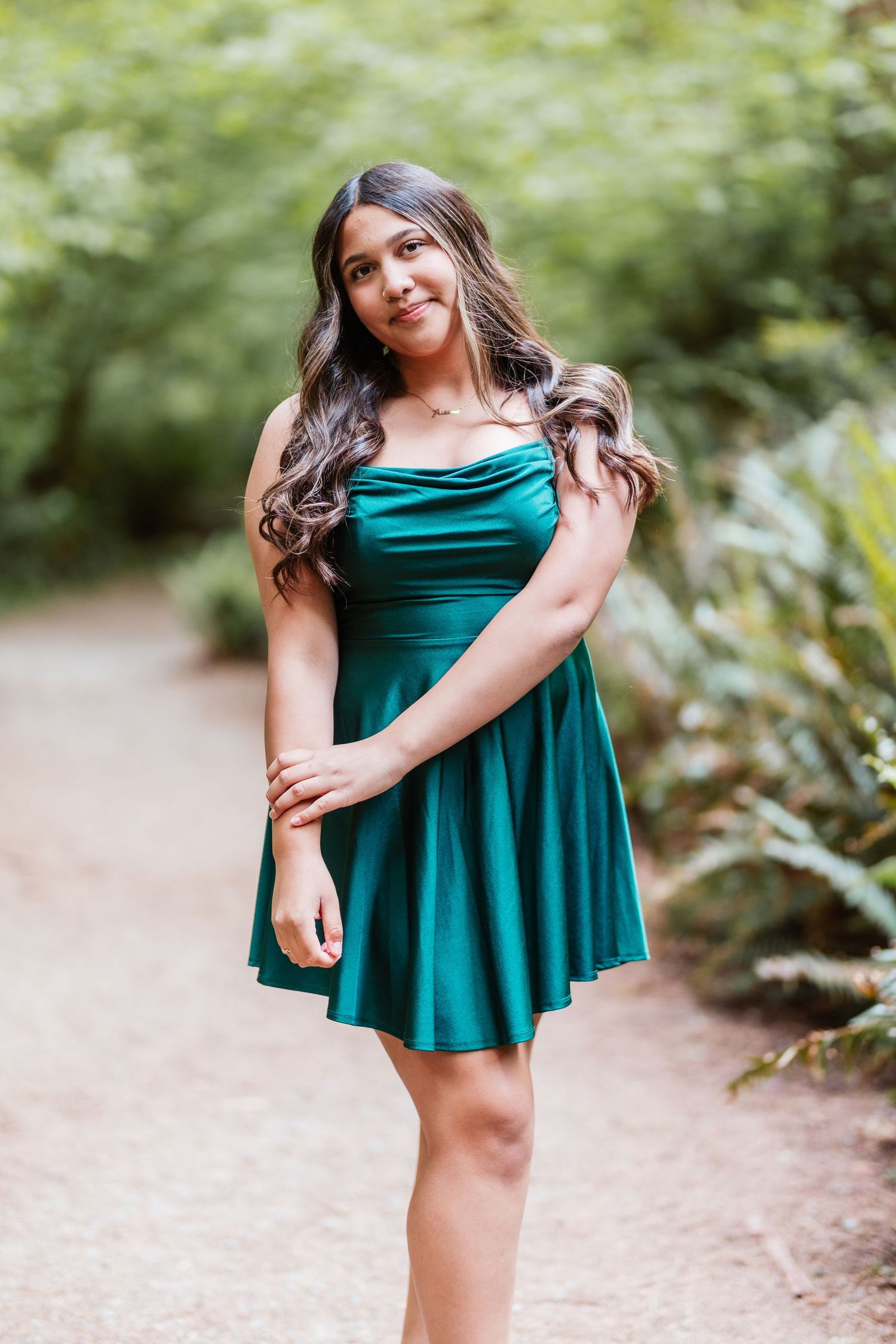 Young woman in teal satin skater dress posing on garden path surrounded by lush green foliage.