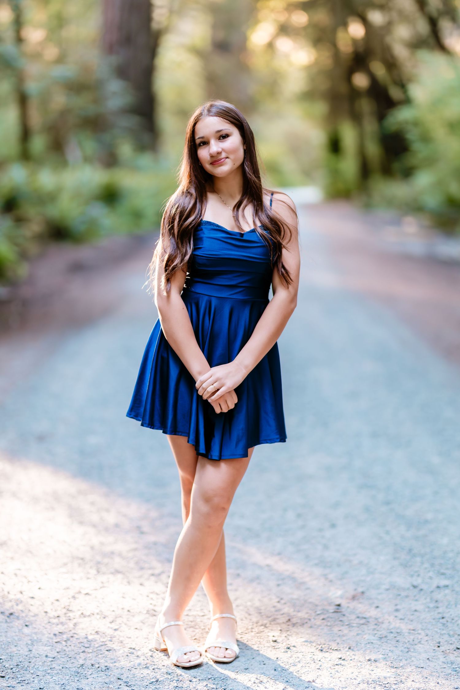 Young woman in royal blue mini dress standing on a tree-lined path, smiling softly.