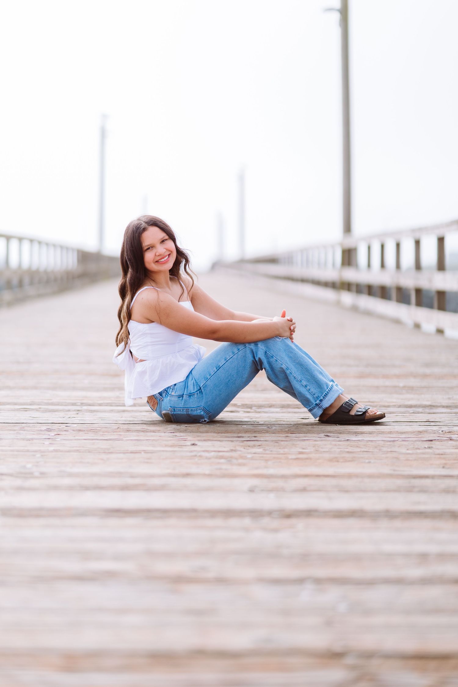 Young woman in white top and jeans sitting on wooden pier, smiling at camera on a calm overcast day.
