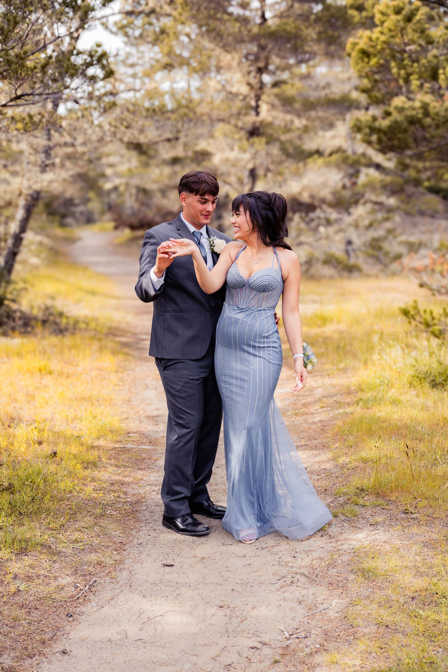 Couple in formal wear dancing on a nature trail, woman in blue gown and man in dark suit, surrounded by trees.