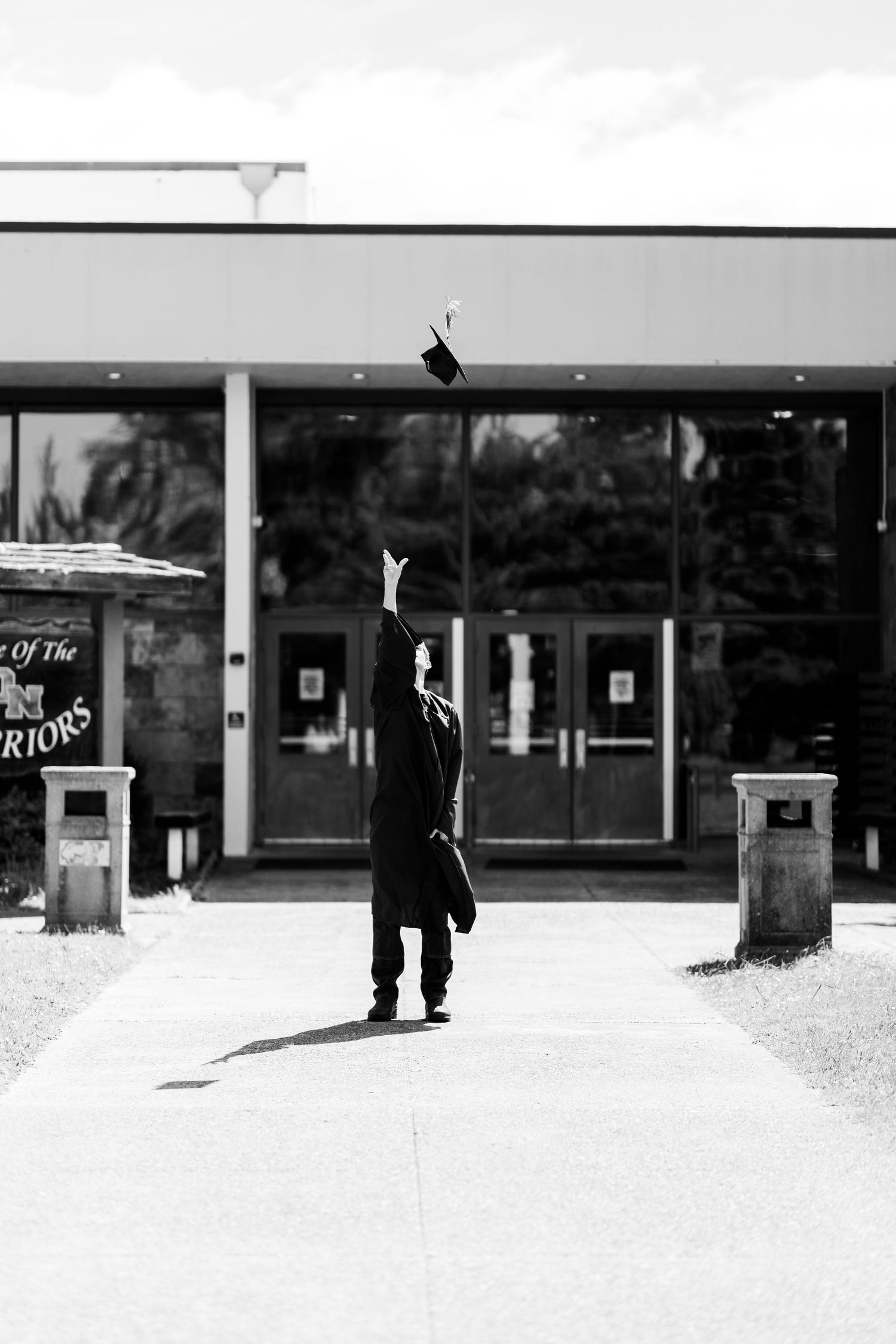 A graduate in cap and gown tosses their mortarboard into the air outside a school building in black and white.