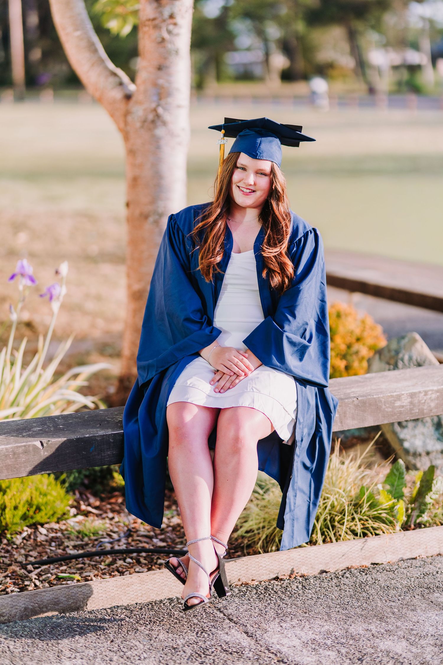 Young woman in blue graduation cap and gown sits on wooden bench outdoors, smiling proudly in white dress.