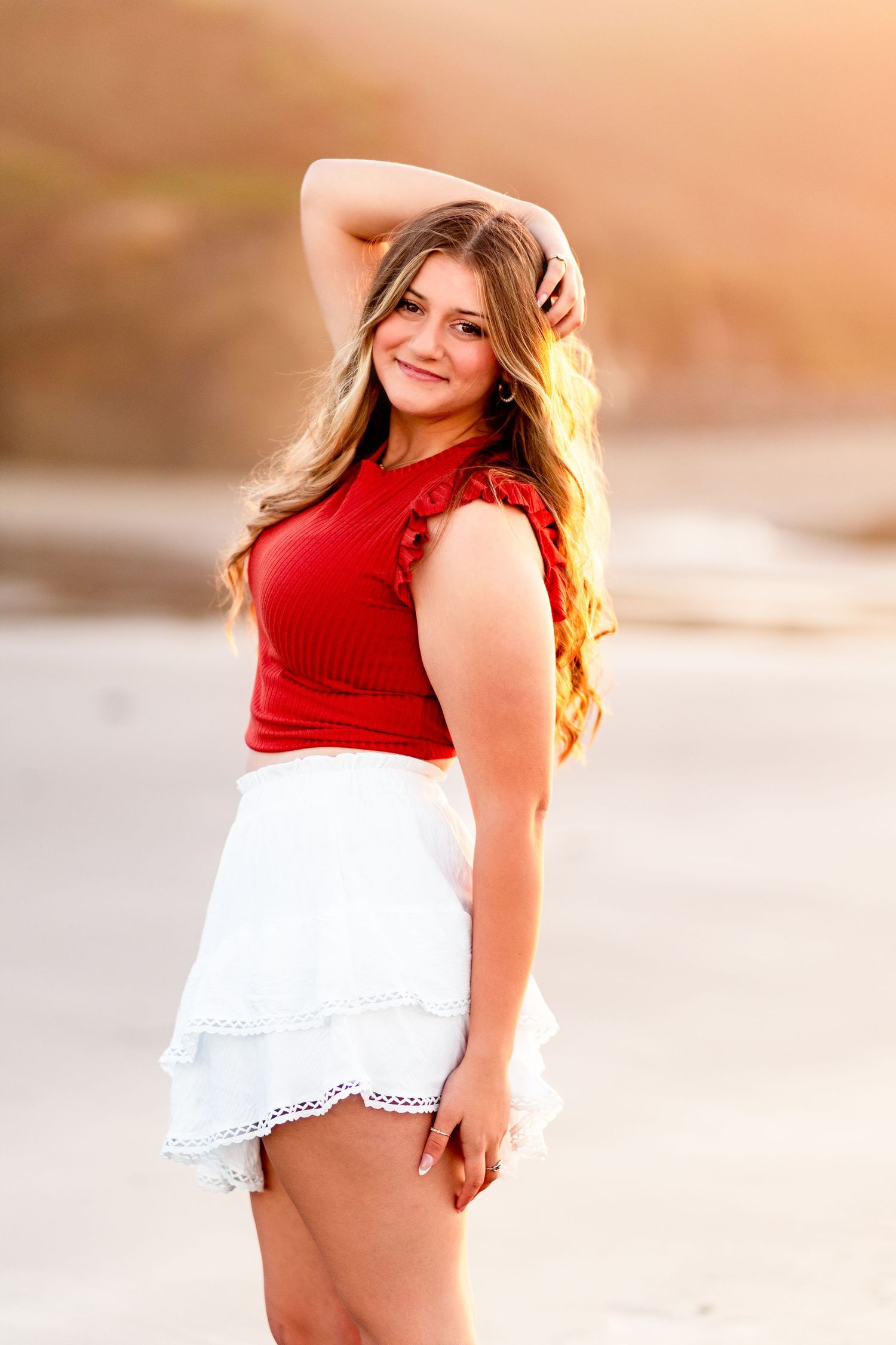 Young woman in red top and white skirt posing on beach at golden hour sunset.