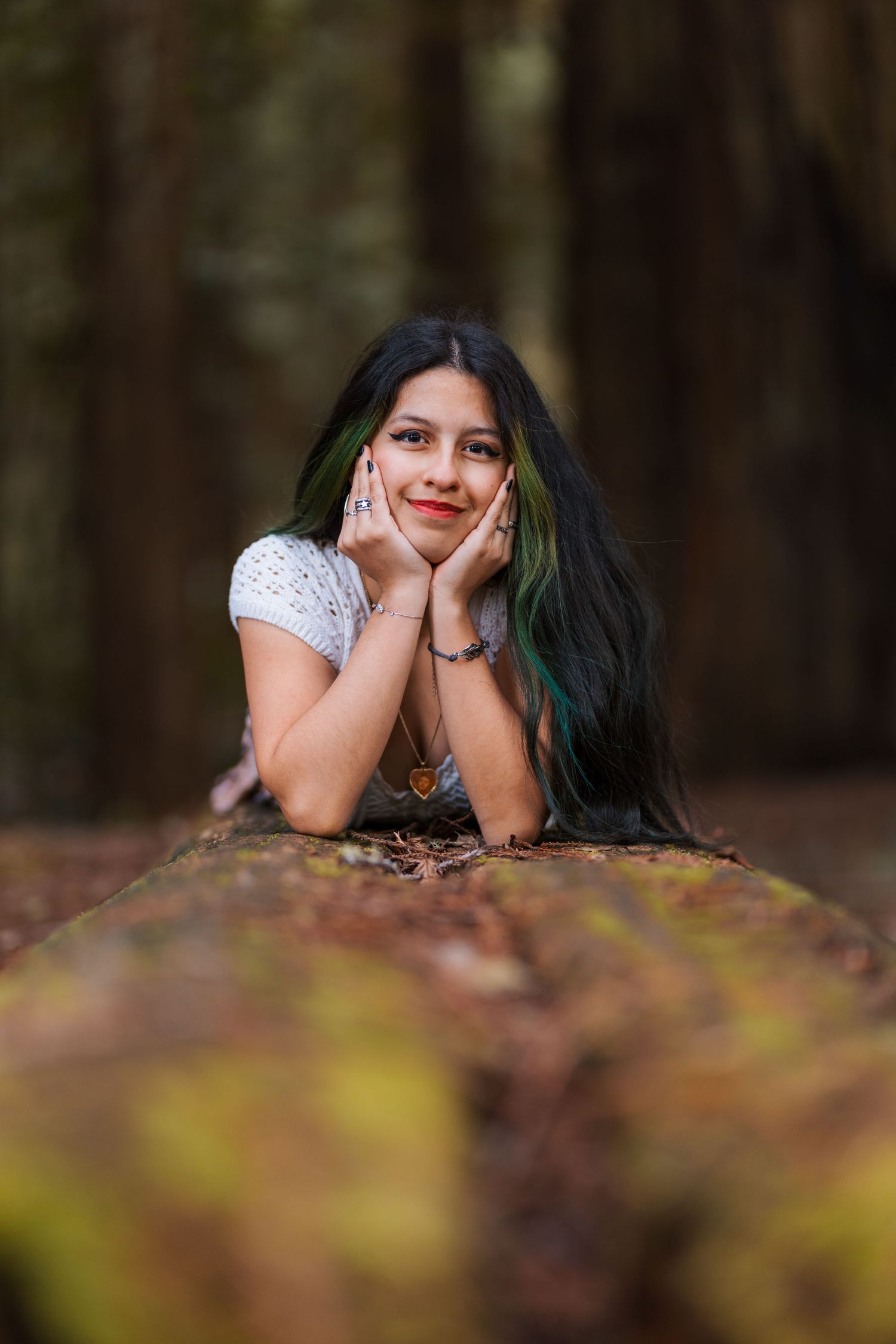 Young woman with long black and green hair rests chin on hands while lying on a mossy log in a redwood forest.