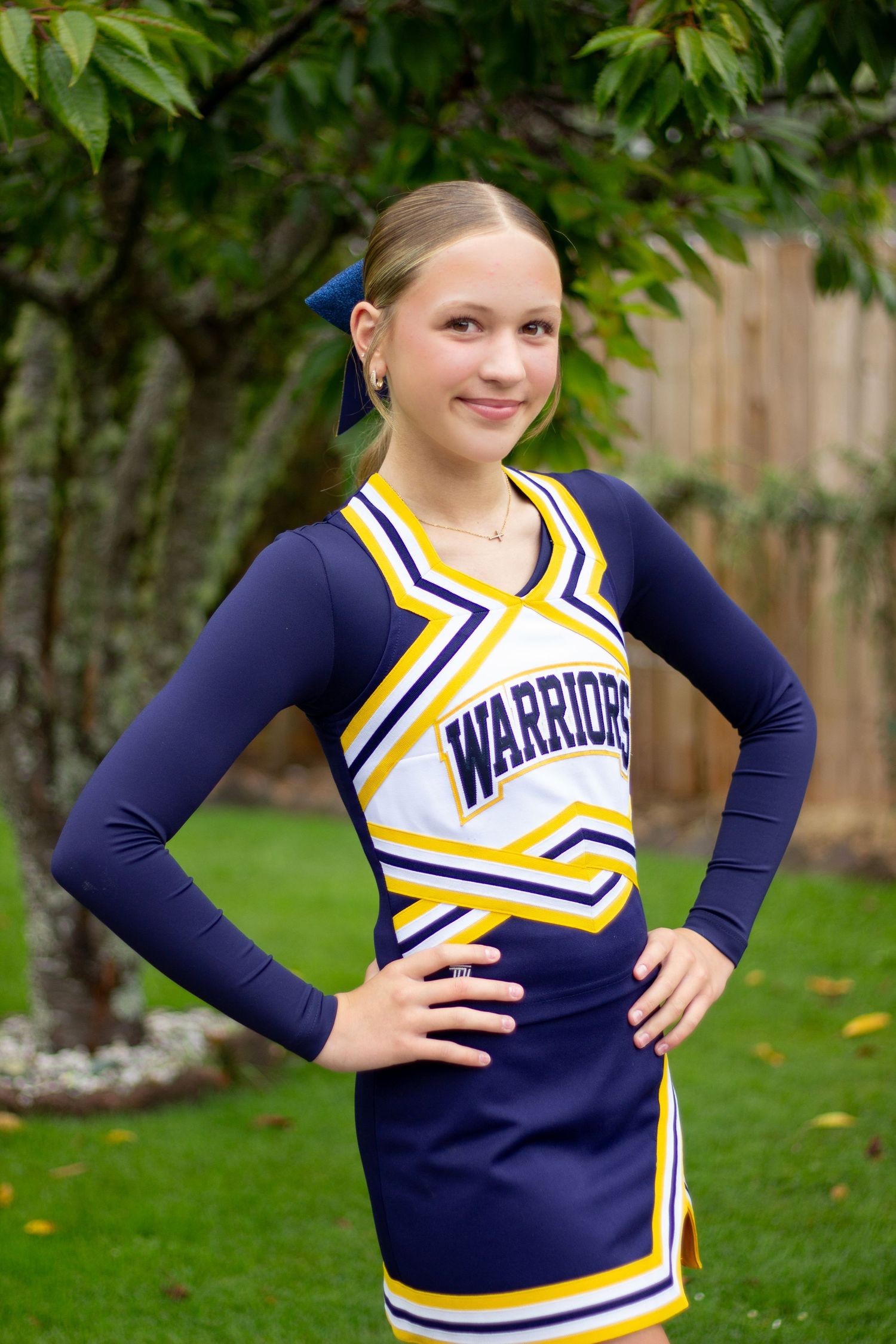 Blonde teenage girl in Warriors cheerleader uniform posing outdoors with hands on hips near green tree.