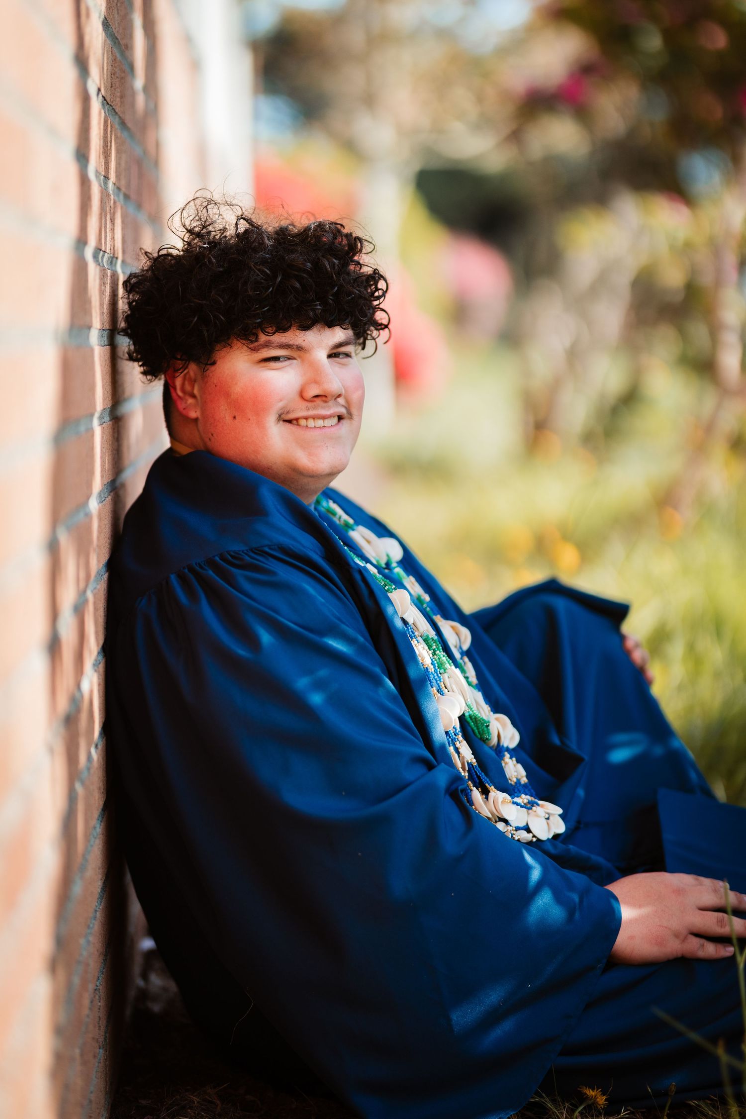 Smiling graduate in blue gown and lei sits against brick wall outdoors on a sunny day.