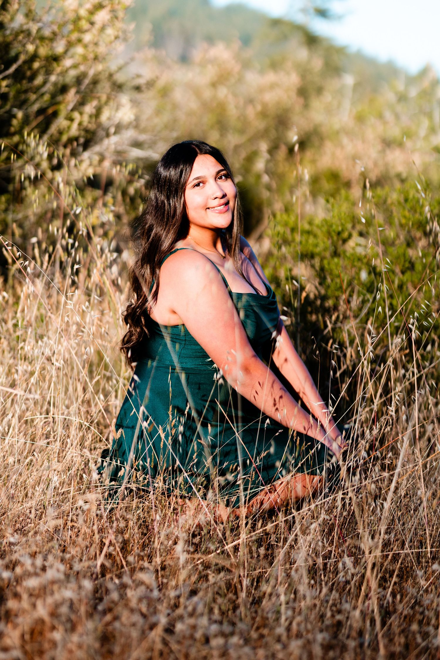 Young woman in green dress sitting in golden dry grass field, smiling outdoors with hills in background.