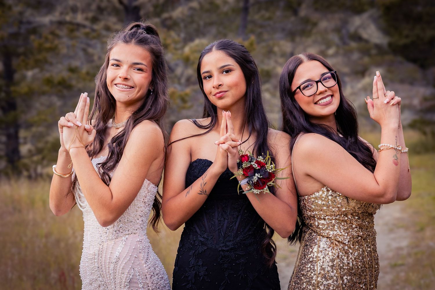Three young women in formal dresses posing outdoors, smiling and making hand gestures at a prom or formal event.