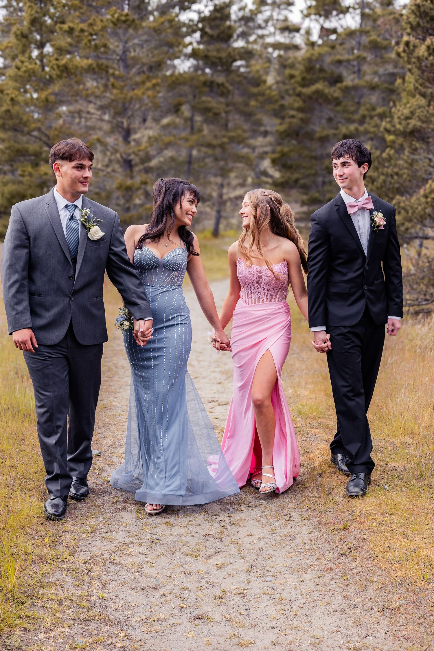 Two couples in prom attire walk hand-in-hand on a dirt path surrounded by trees.