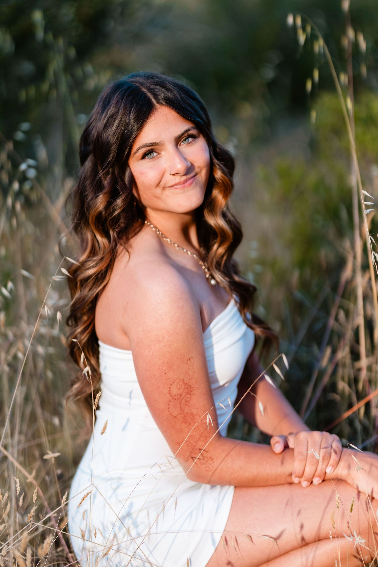 Young woman in white dress with long curly hair sitting in golden field during sunset portrait session.