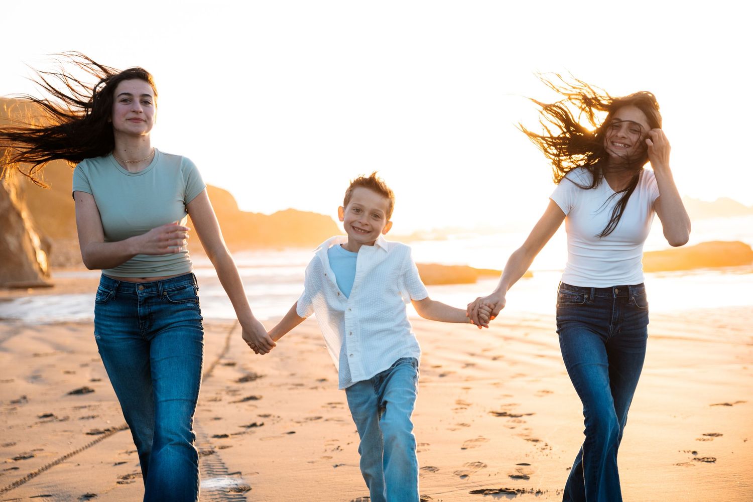 Two women and a young boy holding hands and running on a sunny beach at golden hour.