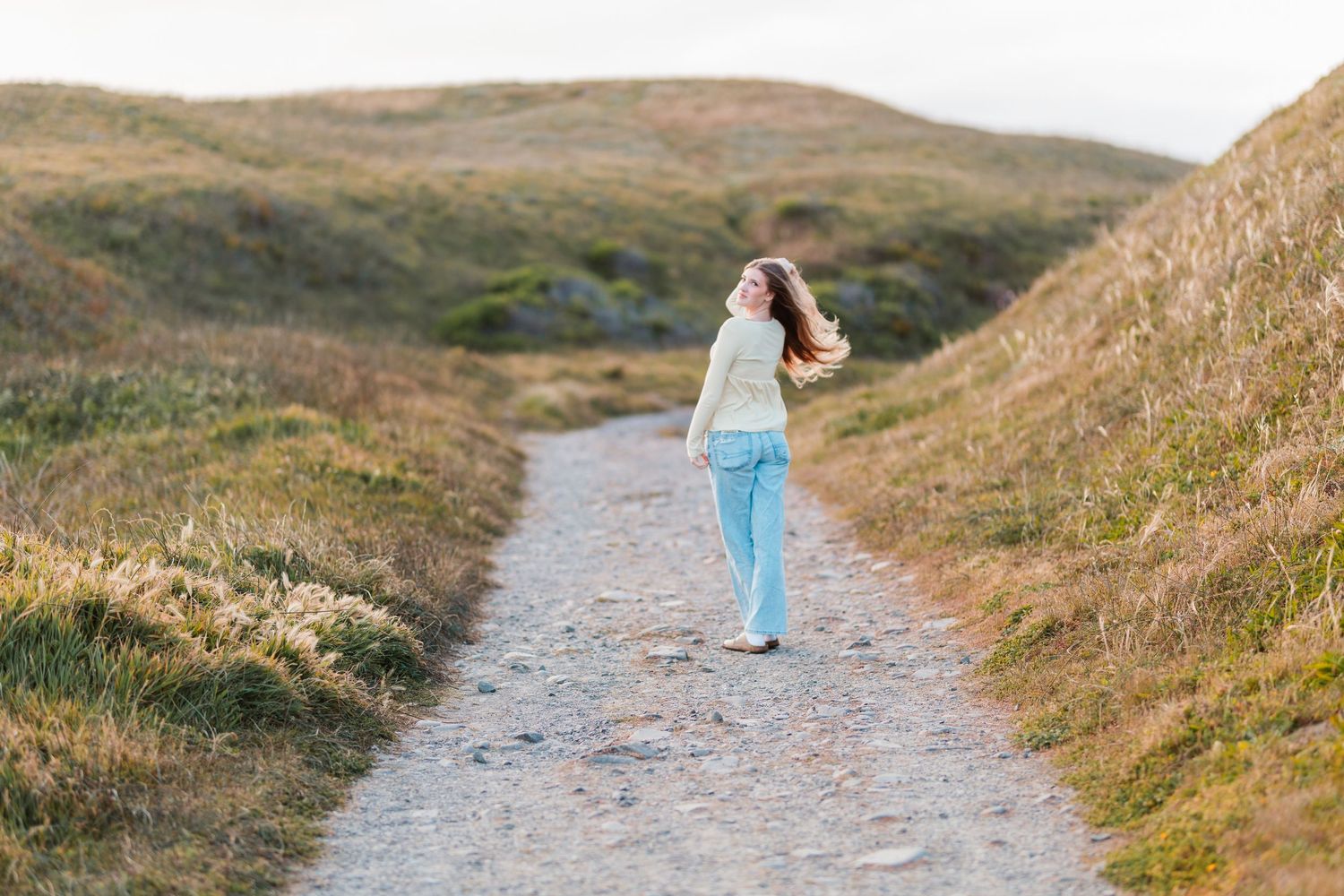 Young woman in yellow top and light jeans walks along scenic path through rolling golden hills.