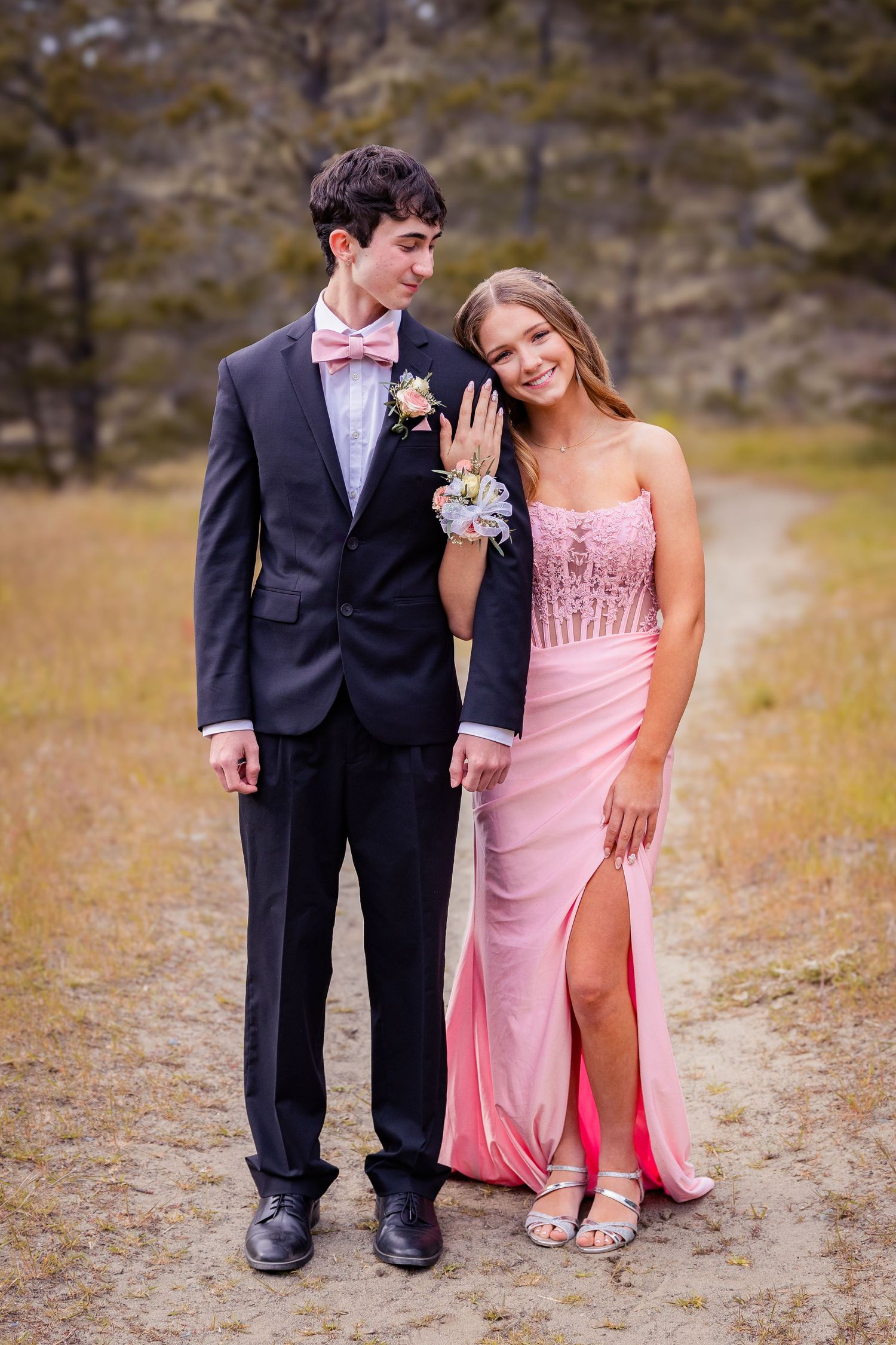 Teen couple in prom attire, boy in black tuxedo with pink bow tie and girl in pink gown, posing outdoors.