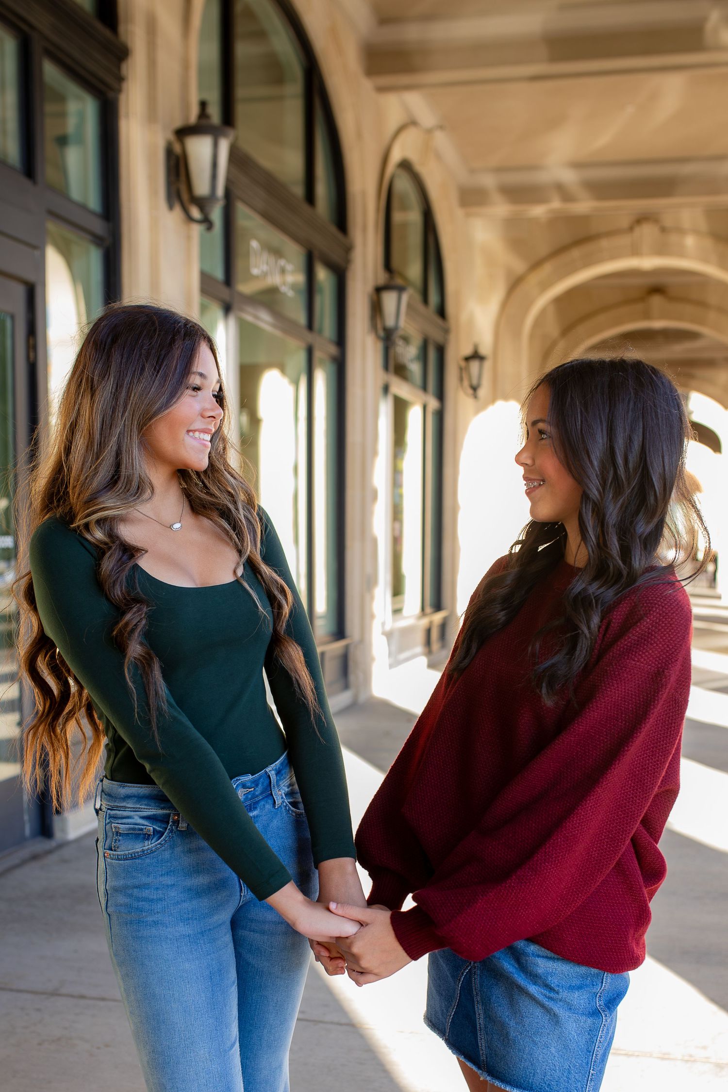 Two friends in casual outfits hold hands and laugh together while standing in a covered outdoor walkway.