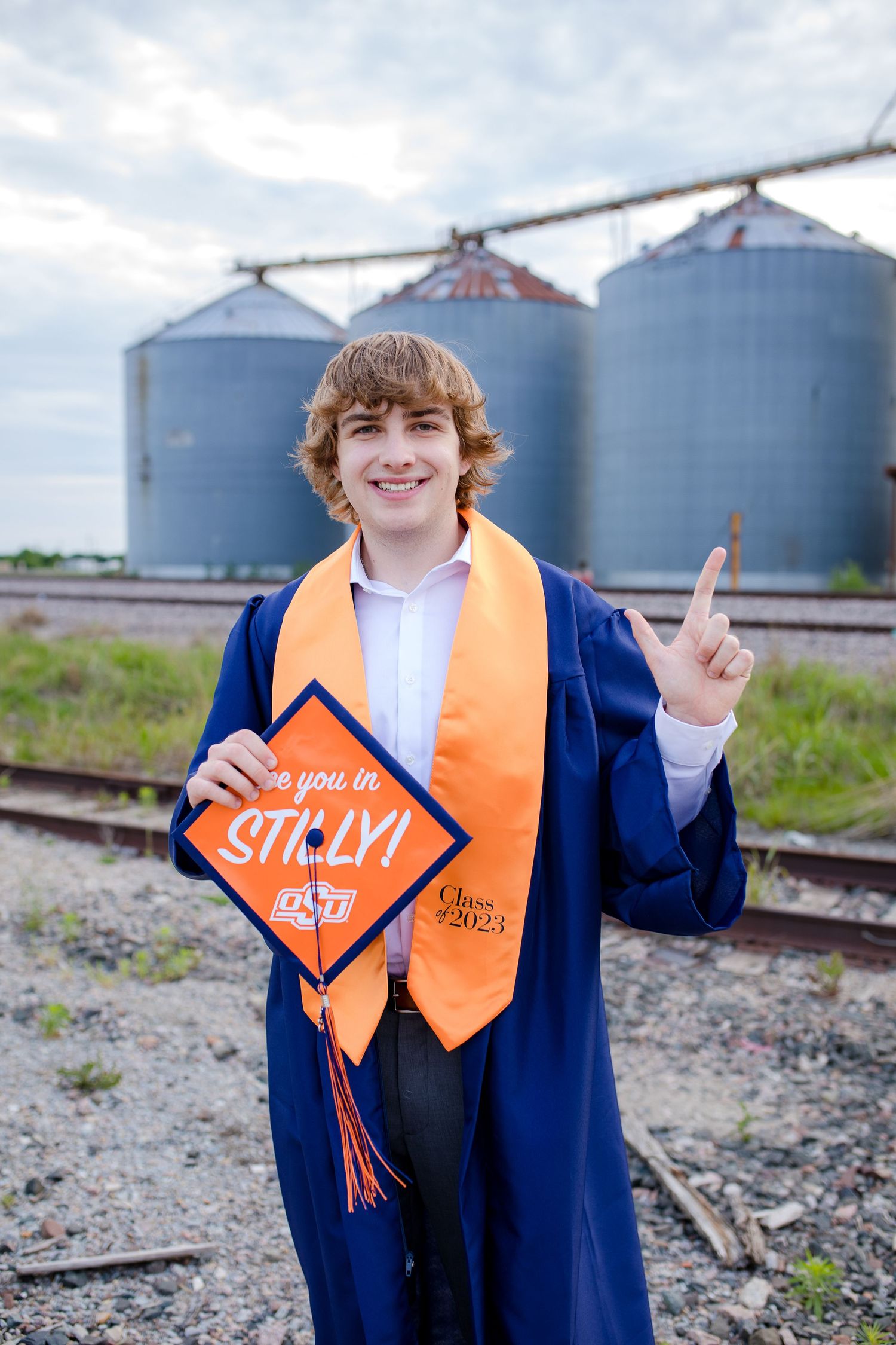 Graduate in blue cap and gown stands in front of grain silos wearing an orange STEM graduation stole with tassel.