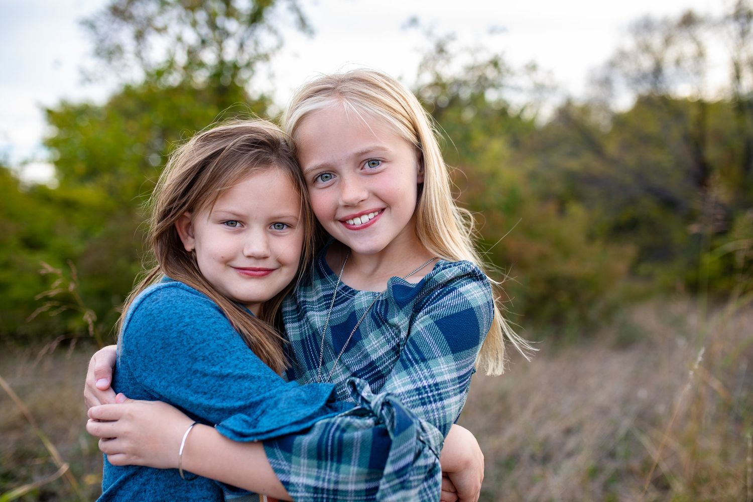 Two young friends in blue clothes share a warm embrace while smiling outdoors in a natural setting.