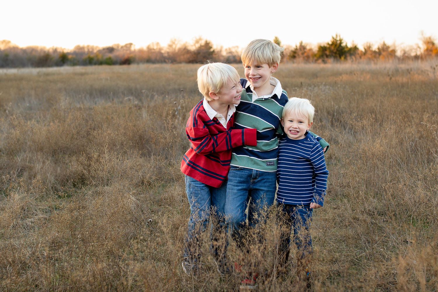 Three children stand together smiling in a tall grass field during golden hour at sunset.