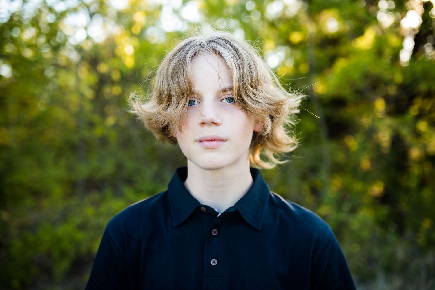Person with wavy blonde hair wearing a black polo shirt stands in front of green foliage in soft natural light.