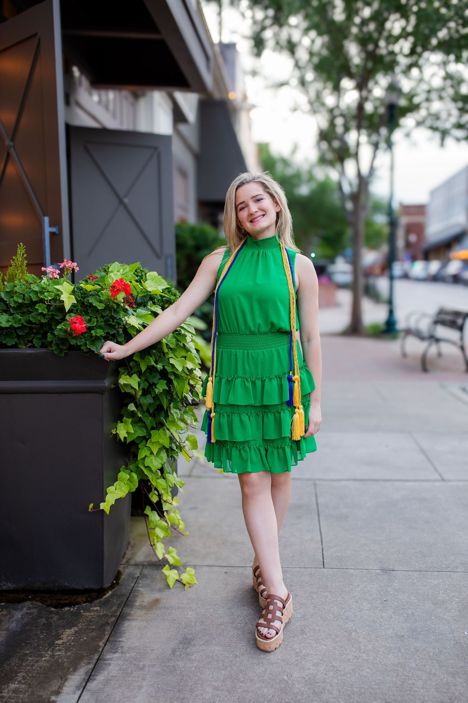 A bright green tiered dress is worn with brown sandals on a sidewalk near decorative planter boxes and storefronts.