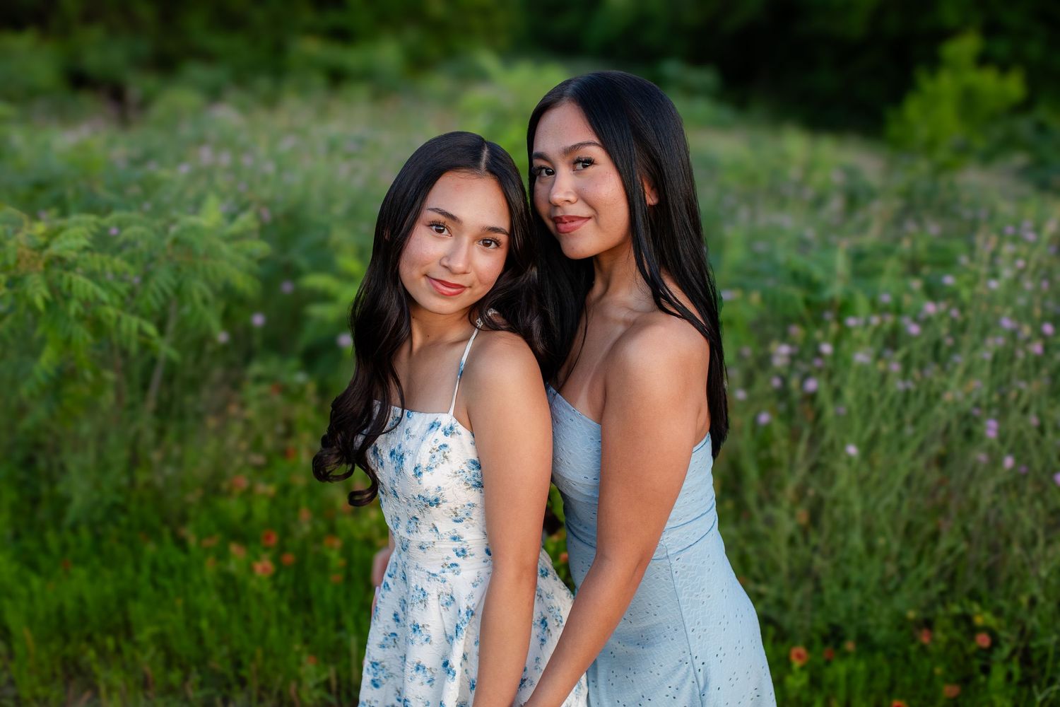 Two people in floral and blue dresses pose together outdoors in a natural meadow setting.