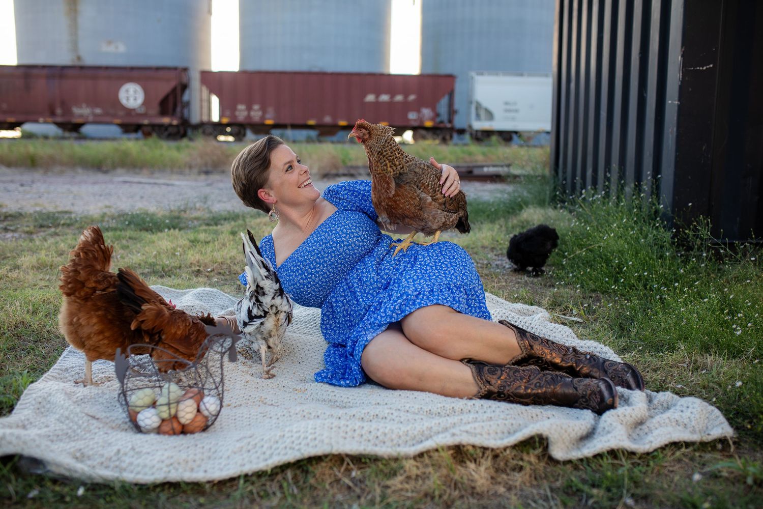 A person lying on a blanket outdoors while playing with a golden-colored dog and chicken in a relaxed setting.