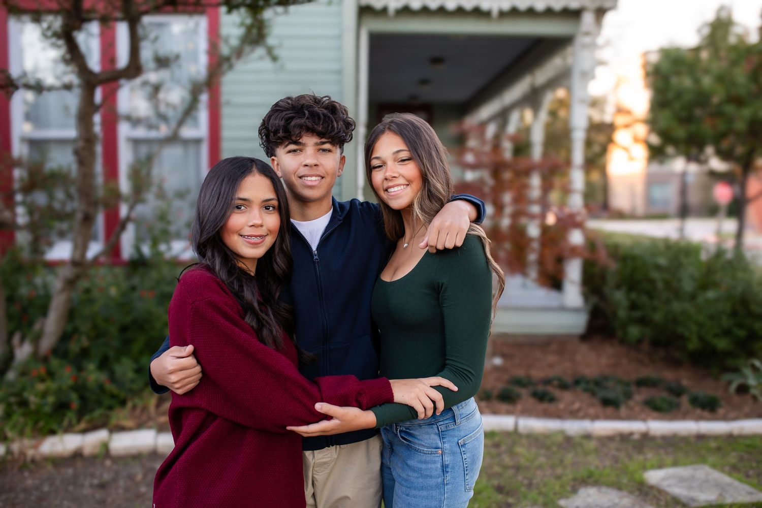 A group of three poses together outdoors wearing casual fall clothing in front of a building at sunset.