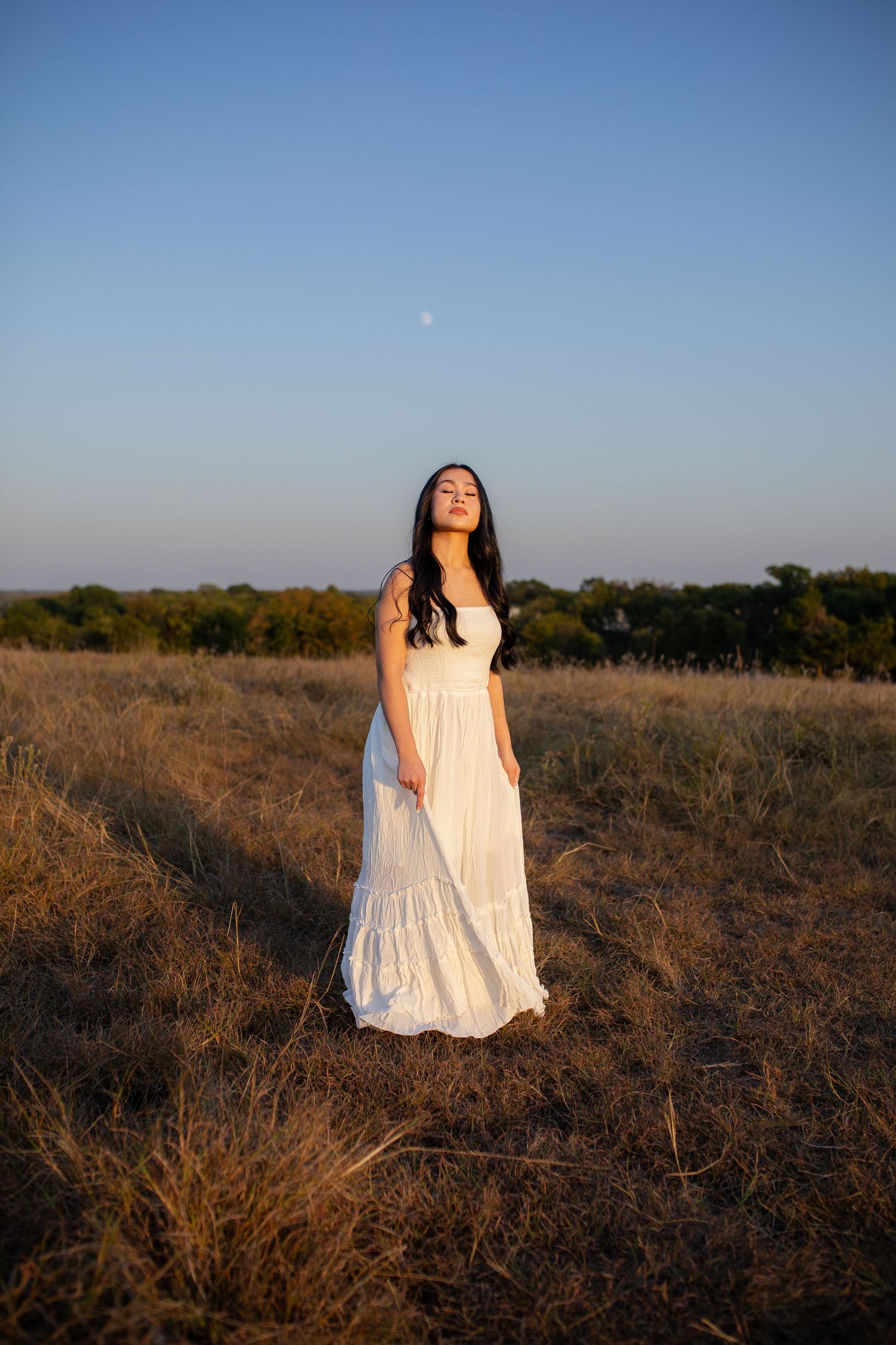 A person in a flowing white dress stands in a golden field at sunset with a faint moon visible in the clear blue sky.