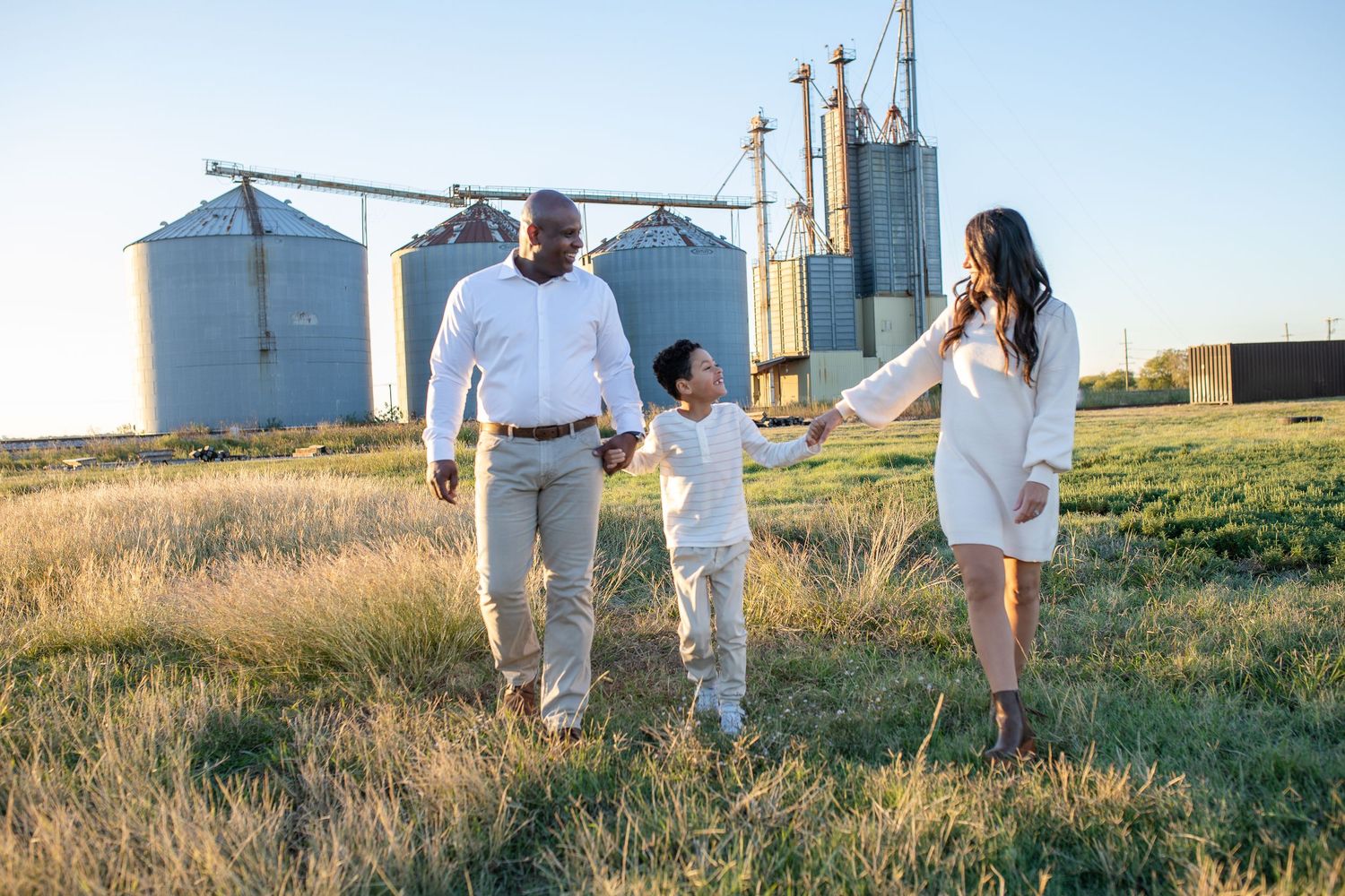 A family walking hand in hand through a grassy field with large grain silos in the background.